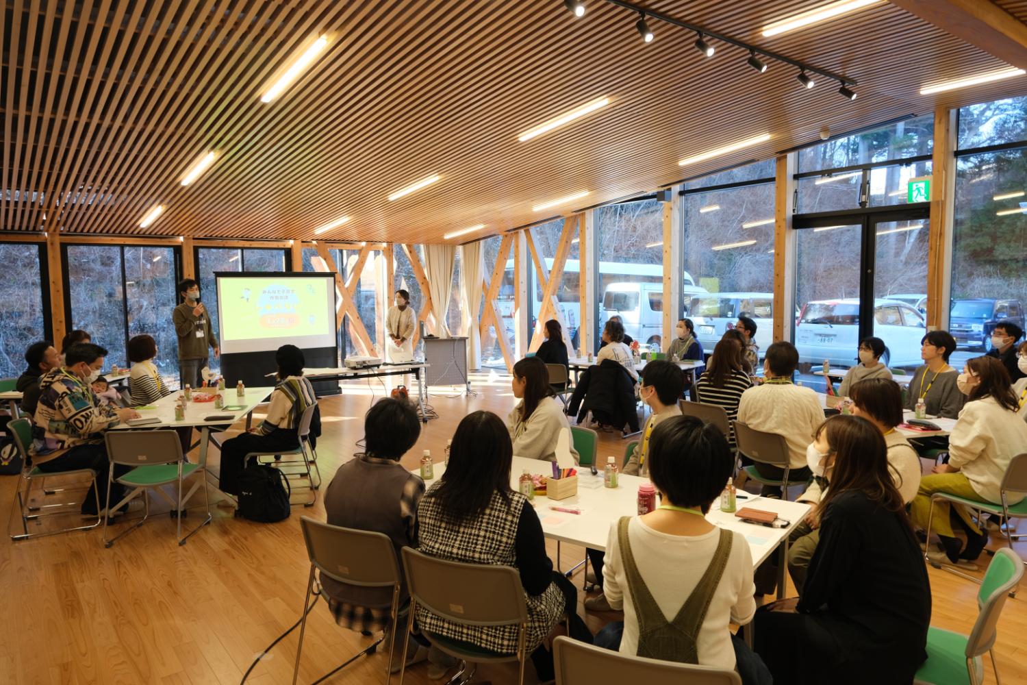 木製の天井と床に囲まれた明るい室内の中央にスクリーンが設置され、登壇者が発表を行うなか、複数のテーブルに分かれて座った参加者たちが話に耳を傾け、学びや交流が進んでいる地域イベントの写真
