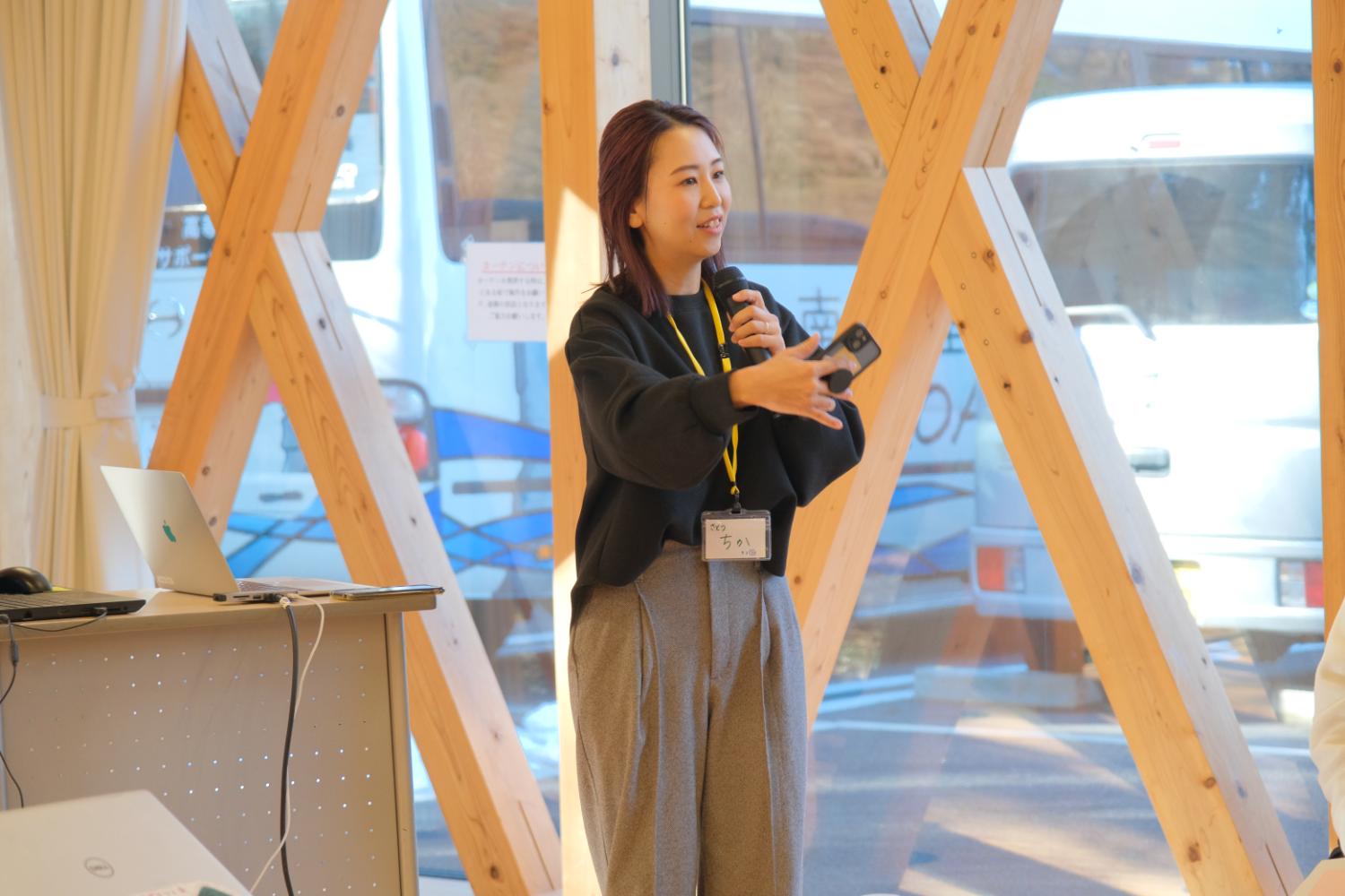 木の梁が交差する大きなガラス窓の前に立つ女性が、マイクとスマートフォンを手に笑顔で話しながら発表をしている写真