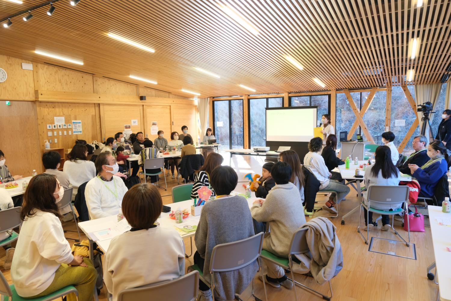 床や壁も明るい木目調で統一された広々とした室内で、複数のテーブルに分かれて座る大人たちが前方のスクリーンと発表者に注目している交流型ワークショップの写真