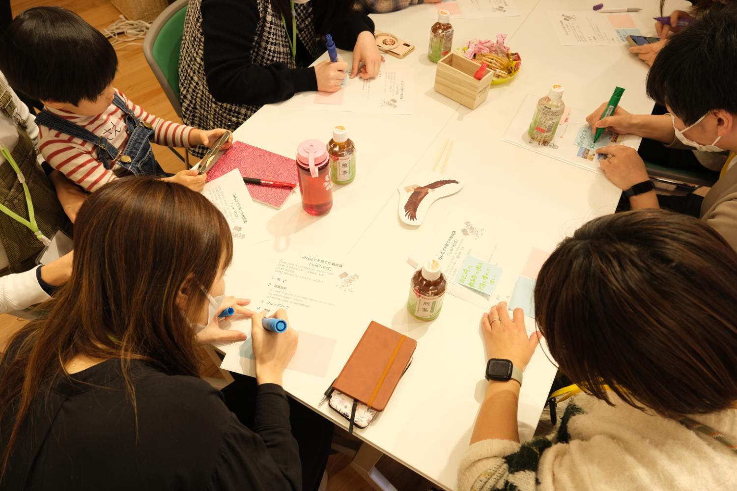 長方形のテーブルを囲んで大人たちがマーカーで紙に何かを書き込んでおり、子どもも1人スマホを手にして参加しているワークショップ中の写真