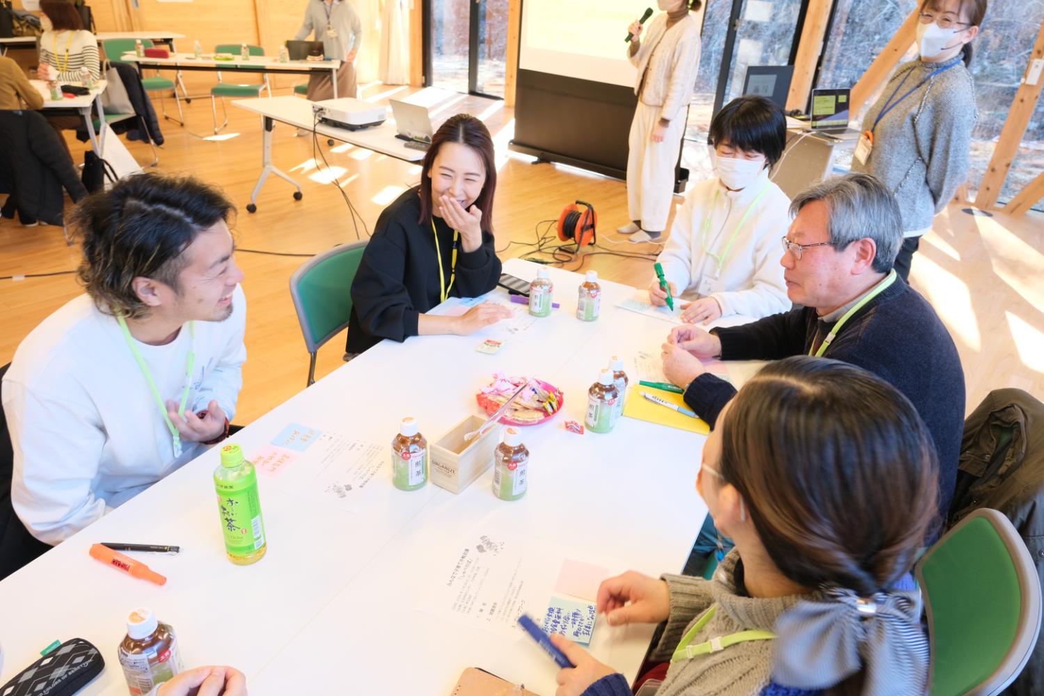 室内で、6人の男女がお茶やお菓子が置かれたテーブルを囲んで話し合いをしており、奥ではマスクを着けた女性がマイクを持って立ち、プロジェクターで発表している地域交流イベントの写真