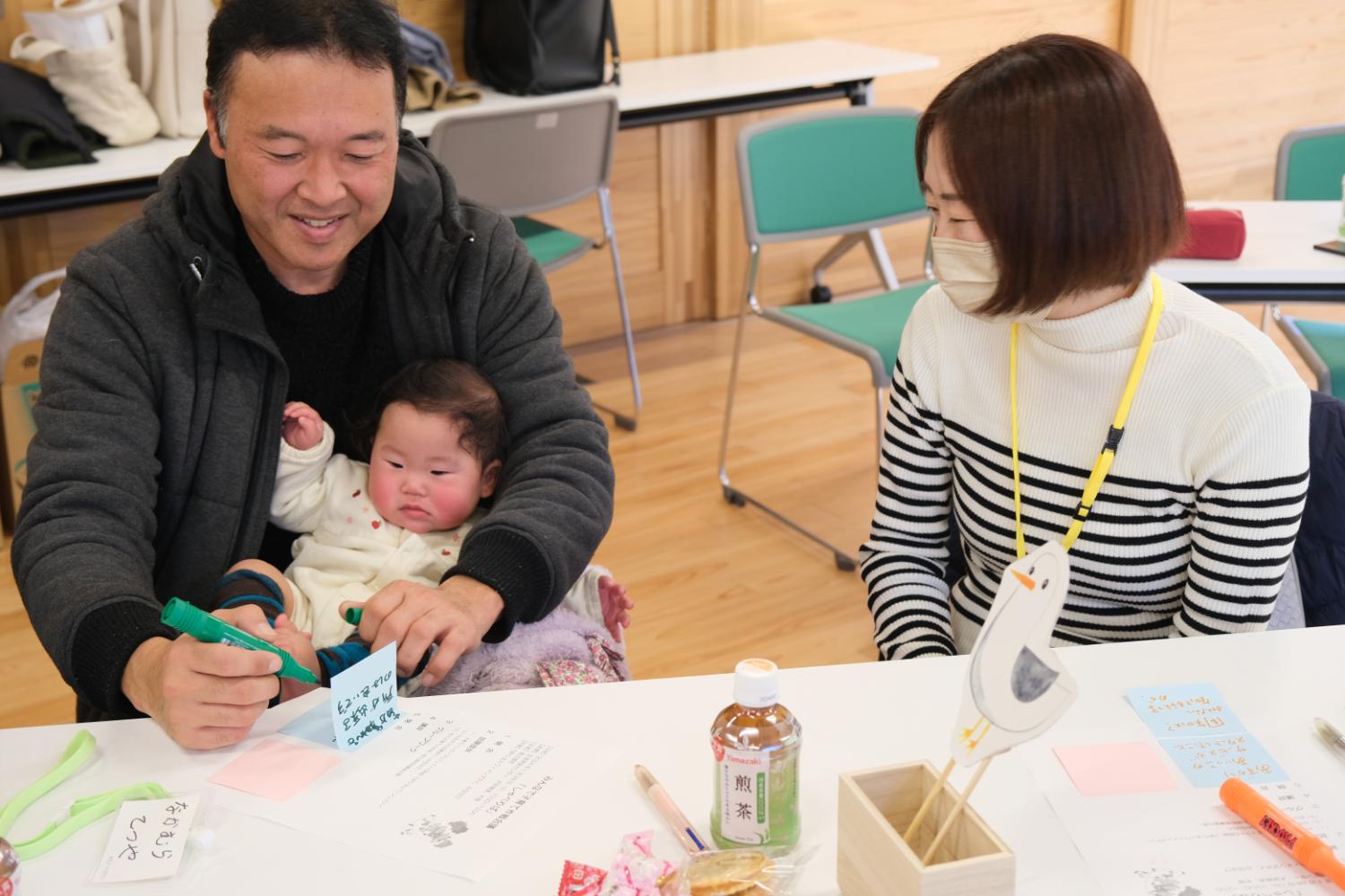 明るい室内で、隣に座るマスク姿の女性が穏やかに見守っている中、赤ちゃんを膝にのせた男性が楽しそうにメモを書いている地域交流ワークショップの写真