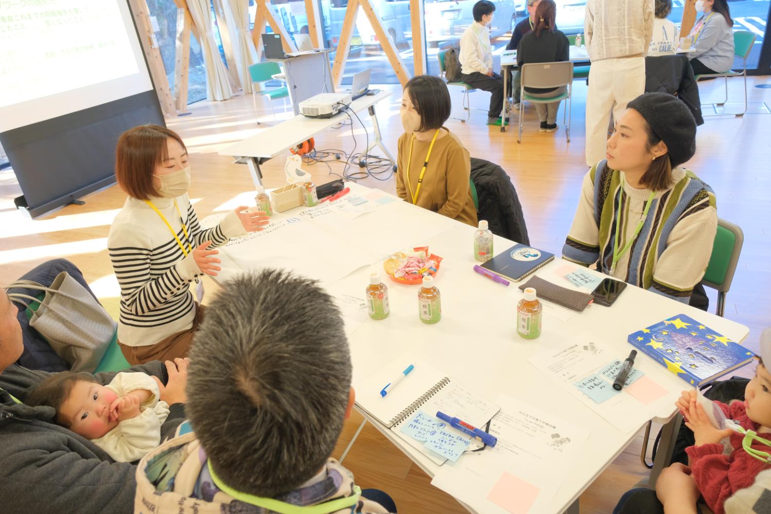 大きな窓から光が差し込む明るい部屋で、大人と子どもが一緒にテーブルを囲んでメモ用紙を見ながら、意見交換が行われているワークショップの写真