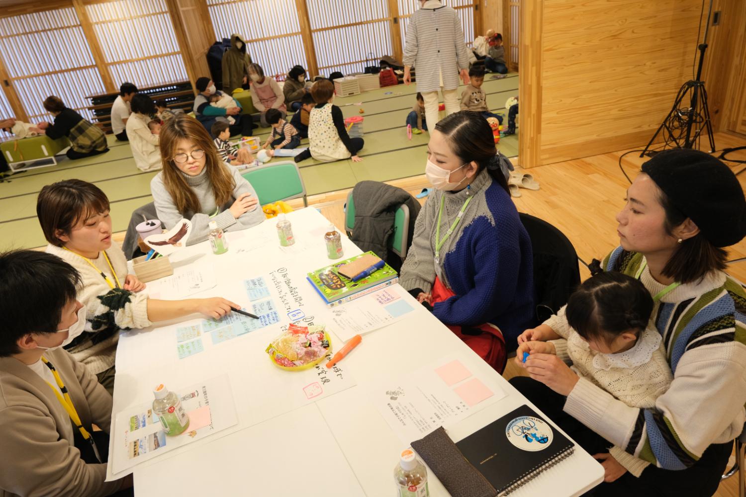 室内で、大人たちがテーブルを囲んで話し合いながら紙にメモを書いており、横の畳スペースでは子どもたちを保護者が見守っている、親子参加型ワークショップの様子の写真