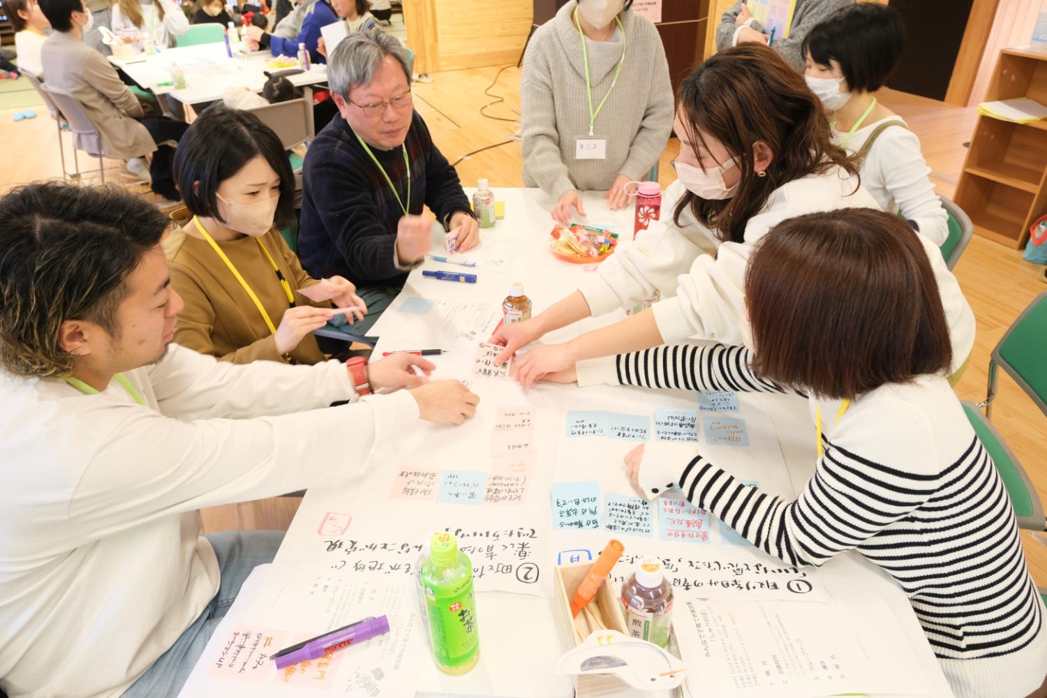 明るい室内で、6人の大人がテーブルを囲み、カラフルな付箋に書かれた意見を並べながら話し合いを進めている、ワークショップの写真