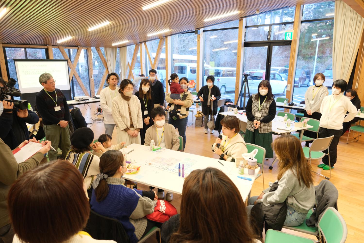 広い部屋で、テーブルを囲んで座る人たちの前でマイクを持った女性が話しており、そのまわりを立って聞く大人たちや、子どもを抱いた人、カメラを構える人もいるにぎやかなワークショップの写真