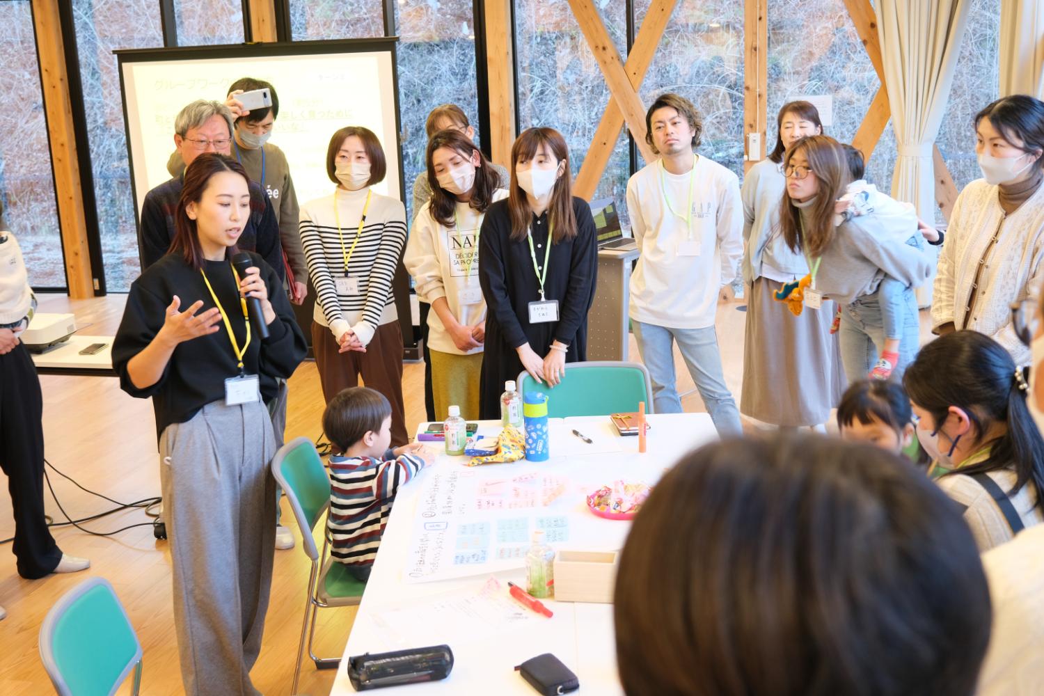 明るい部屋で、子どもが座るテーブルを囲んでマスクをした参加者や子どもを背負った女性が立ち、黒い服の女性がマイクを持って話していることを真剣な表情で聞いているワークショップ発表の写真