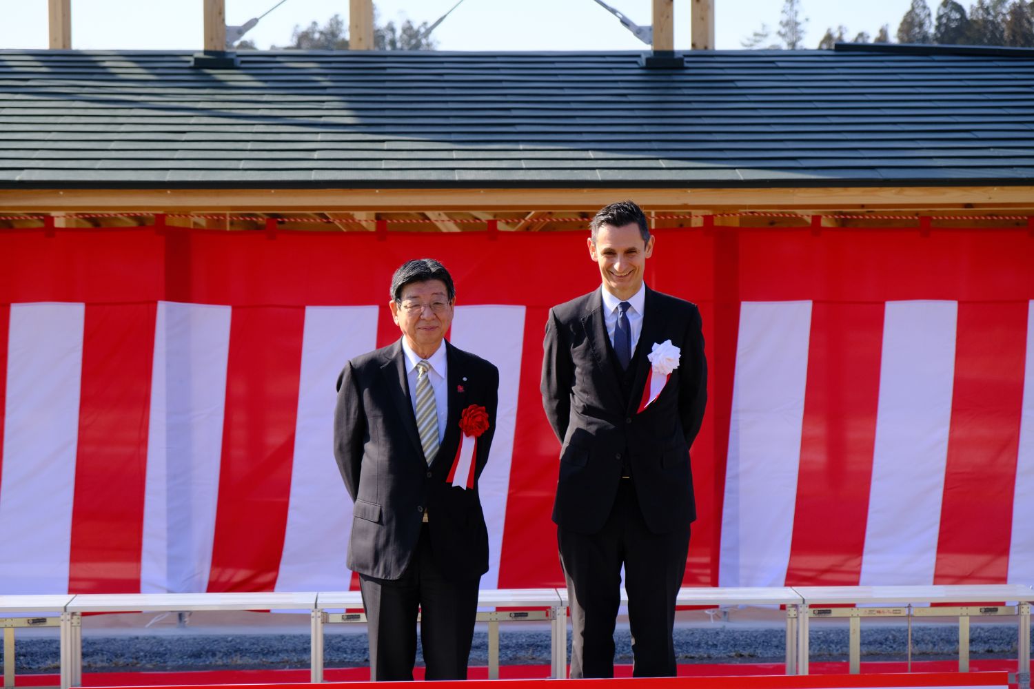 紅白幕が張られた式典会場で、左胸に胸章リボンをつけたスーツ姿の佐藤町長と代表の男性が並び記念撮影している写真