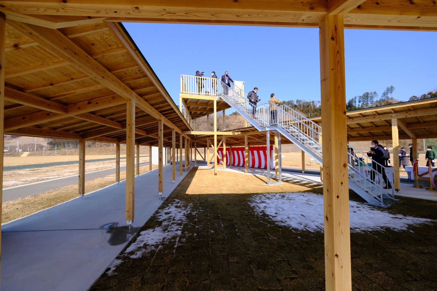 地面に雪が残った広場に設置された、数人の報道関係者が撮影している階段付き展望台を、木製の屋根付き通路から撮影した写真