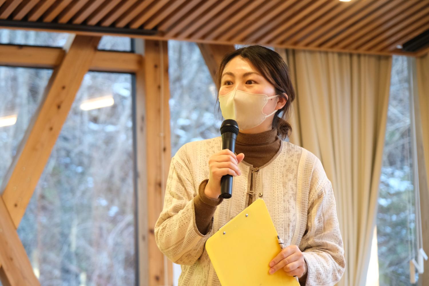 室内で、白いマスクとベージュ系の服を着た女性がマイクと黄色いバインダーを持って話している、地域イベント会場での発表の写真
