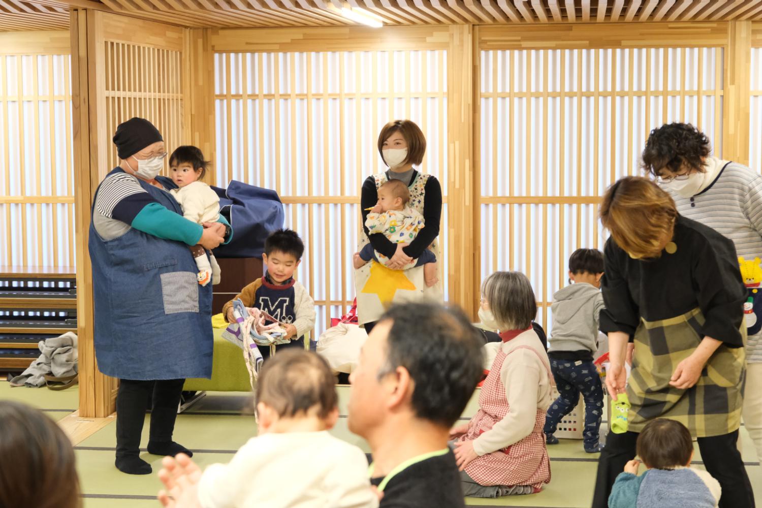 和室のような空間で、大人たちが赤ちゃんや幼い子どもたちの世話をしながら、畳の上でくつろいだり遊んだりしており、エプロンを着た大人が抱っこや着替えを手伝っている子育支援活動の写真