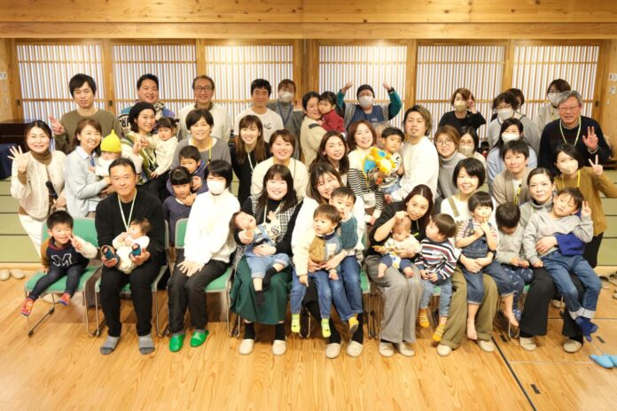 大人と子ども合わせて40人以上が二列に並び、笑顔でカメラに向かってポーズをとり、前列では多くの保護者が子どもを抱っこして座り、後列では多くの人が立っており、奥には縦格子の窓が並ぶ室内での家族交流会の写真