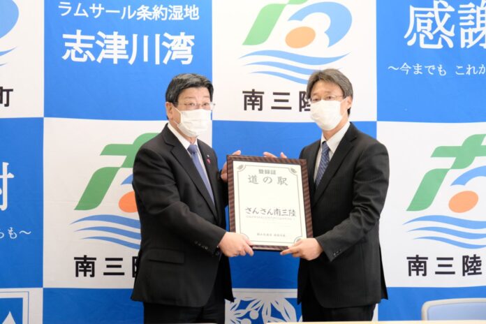 青と白の格子模様のパネルの前にスーツ姿でマスクを着用した二人の男性が「さんさん南三陸」の登録証を持ち記念撮影している写真