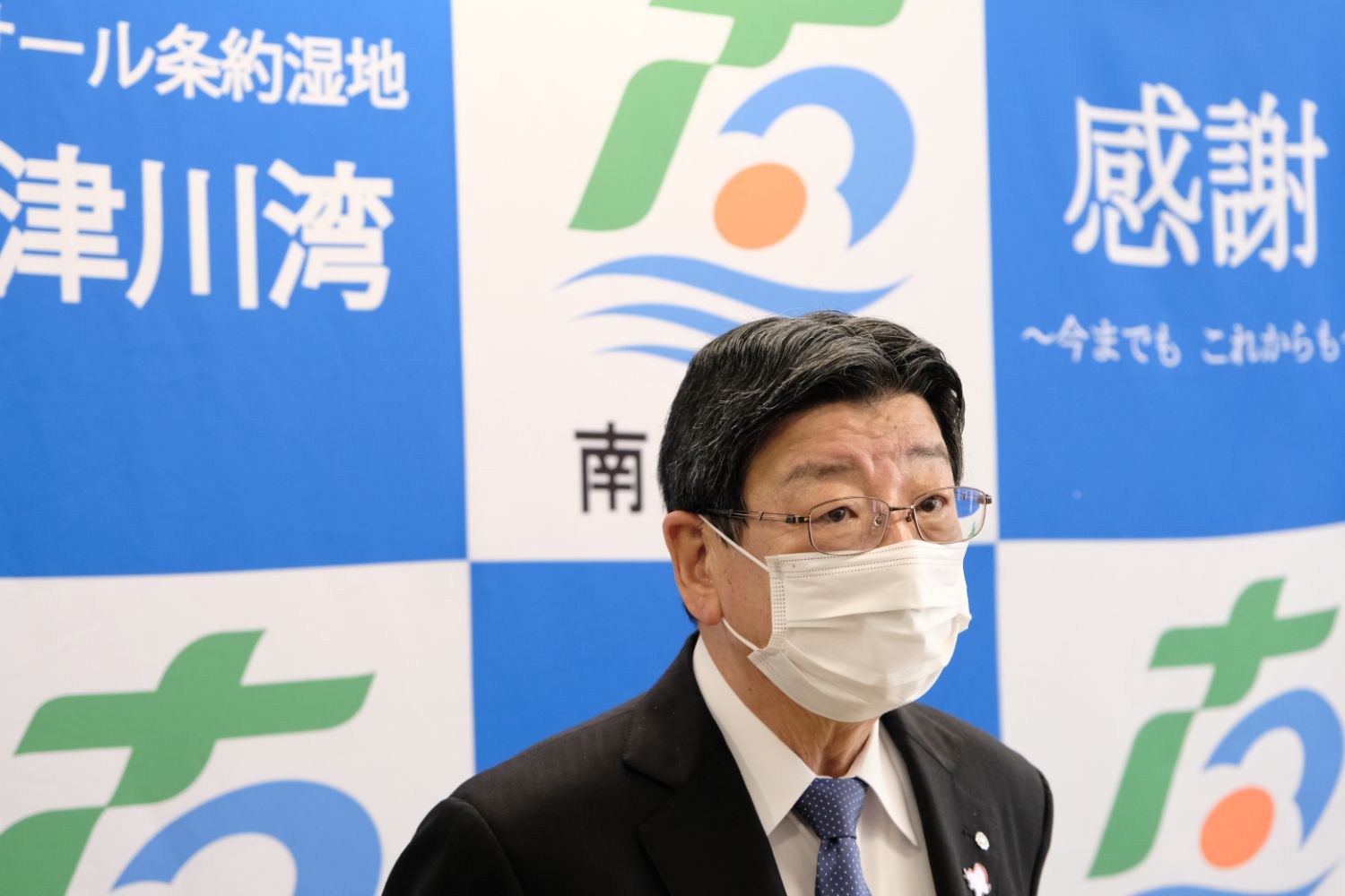 青と白の格子模様のパネルの前にスーツ姿でマスクを着用した男性が話をしている様子の写真