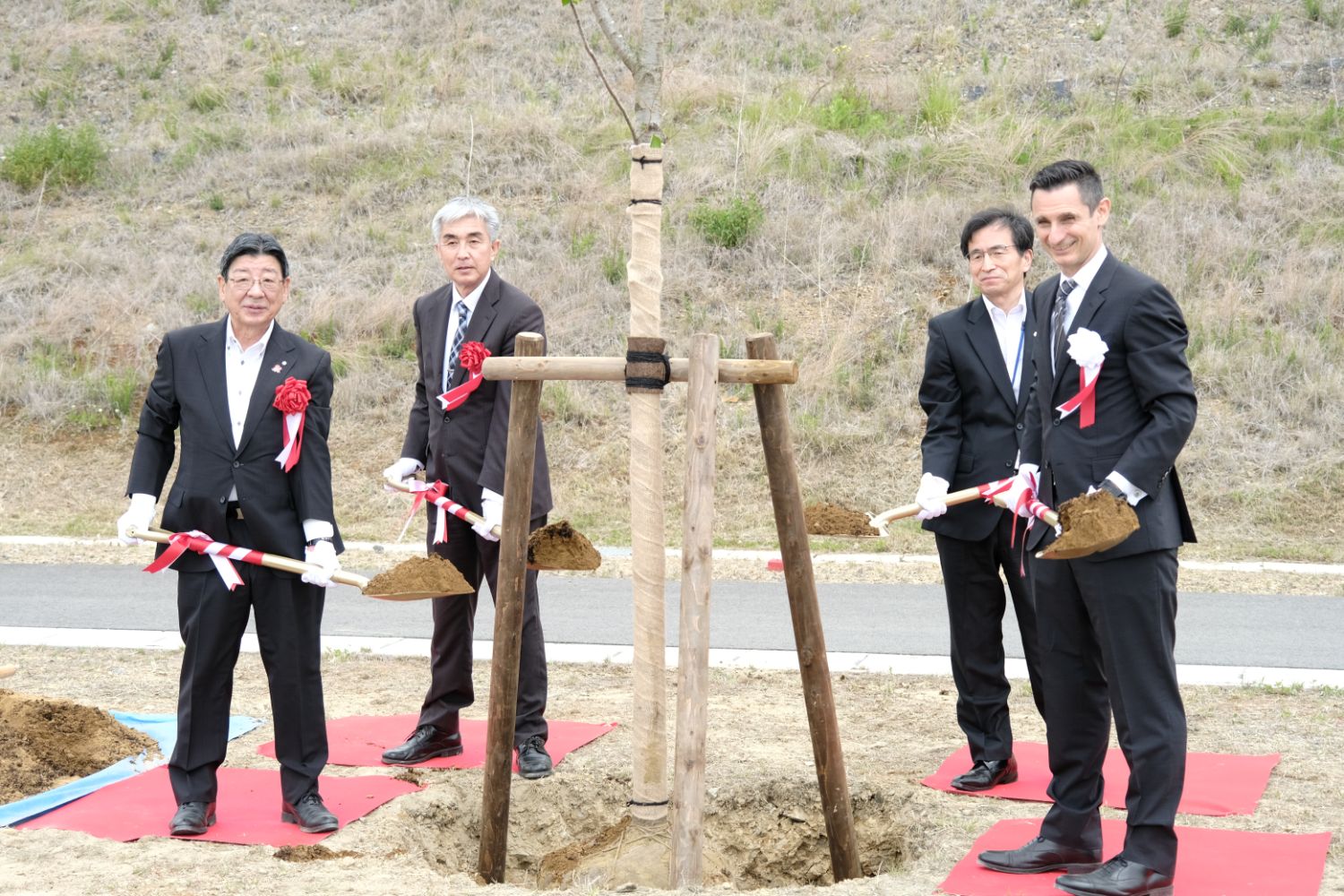 赤い敷物の上に4人のスーツ姿の男性が木の苗を囲んで土をのせた植樹用のスコップを高く持ち上げて記念撮影をしている写真