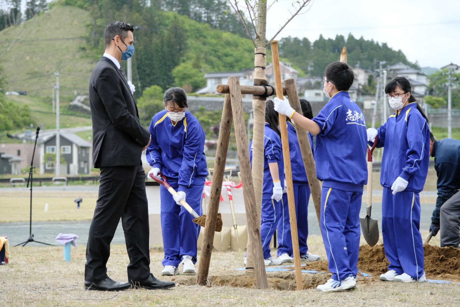 アンドリュー・ガウチ社長がマスクをつけて見守る中、青いジャージの中学生たちが木の苗に土を入れをしている写真