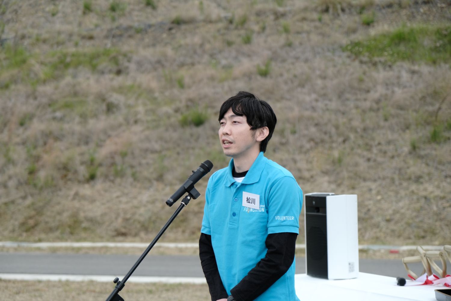 水色のポロシャツを着た男性が屋外でマイクで話している写真