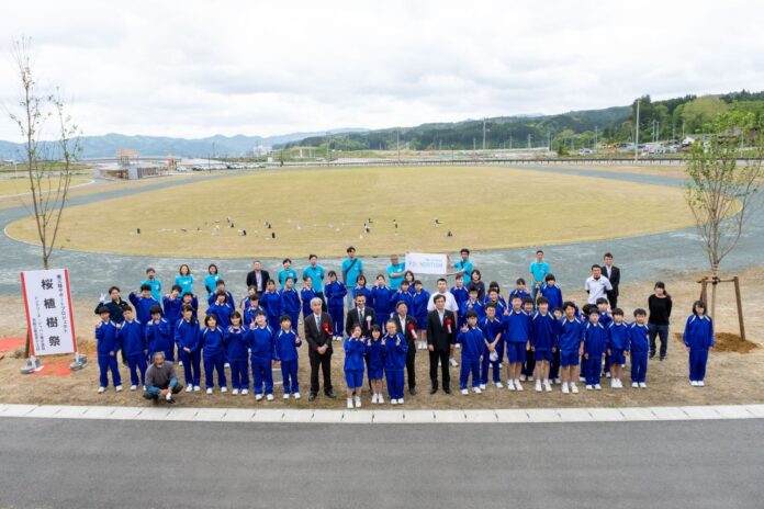 松原公園の広場で青いジャージ姿の多くの生徒と、水色のポロシャツの方や黒いスーツを着た方が「桜植樹祭」と書かれた看板の前で記念撮影をしている写真