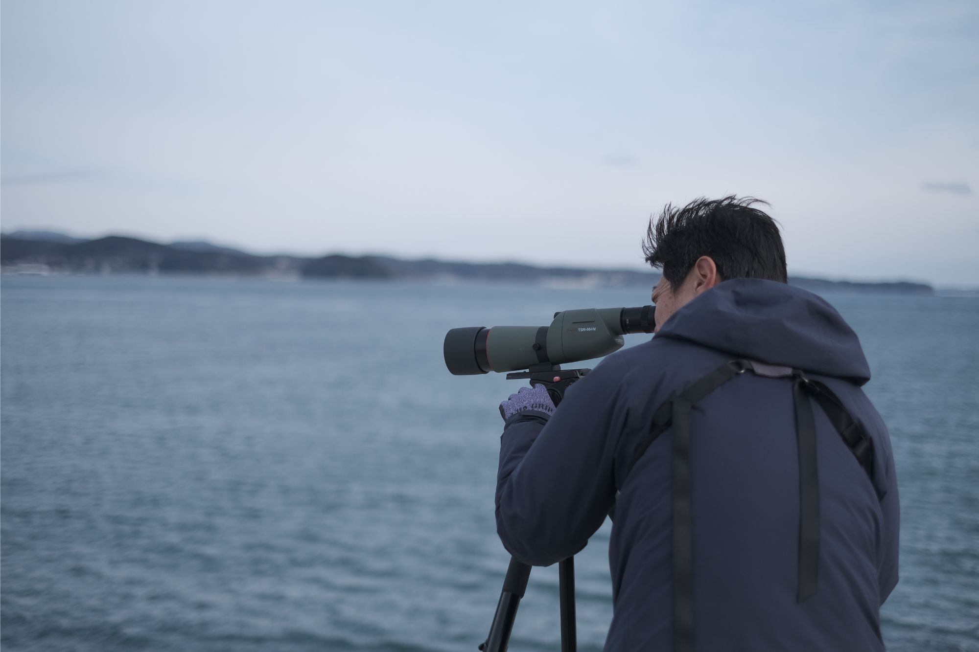 三脚付きの望遠鏡に片目を接眼レンズに当てて観察する鈴木さんの写真