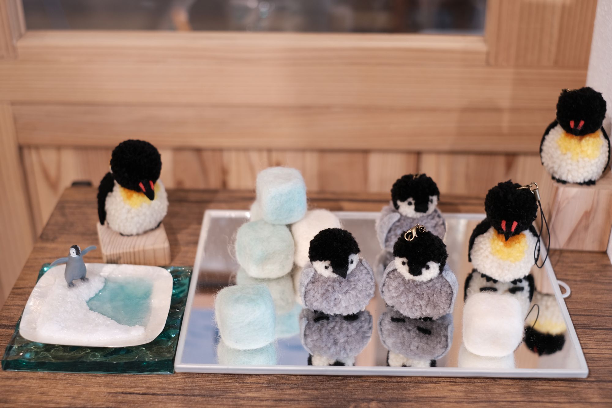 黒い頭と白または灰色の体を持つ6体の手作りペンギン人形と、白や水色のフェルトキューブが反射面の上に並べられ、左側には氷のような台の上に立つ小さなペンギンのミニチュアが水辺のそばに配置されたクラフト作品の写真