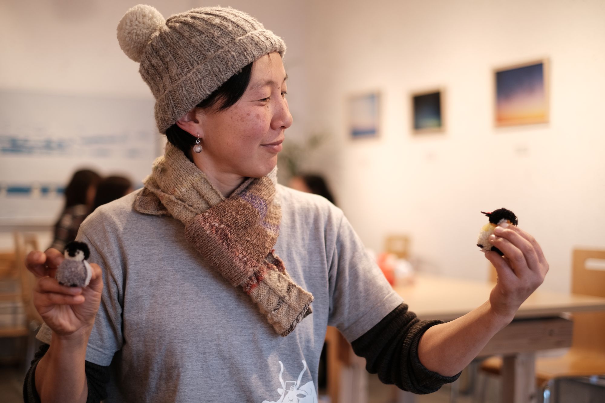 ニット帽とマフラーを身につけた石井洋子さんが、両手に小さなペンギンのぬいぐるみを持っており、背景にはテーブルや椅子、壁に飾られた額縁が見える室内の写真