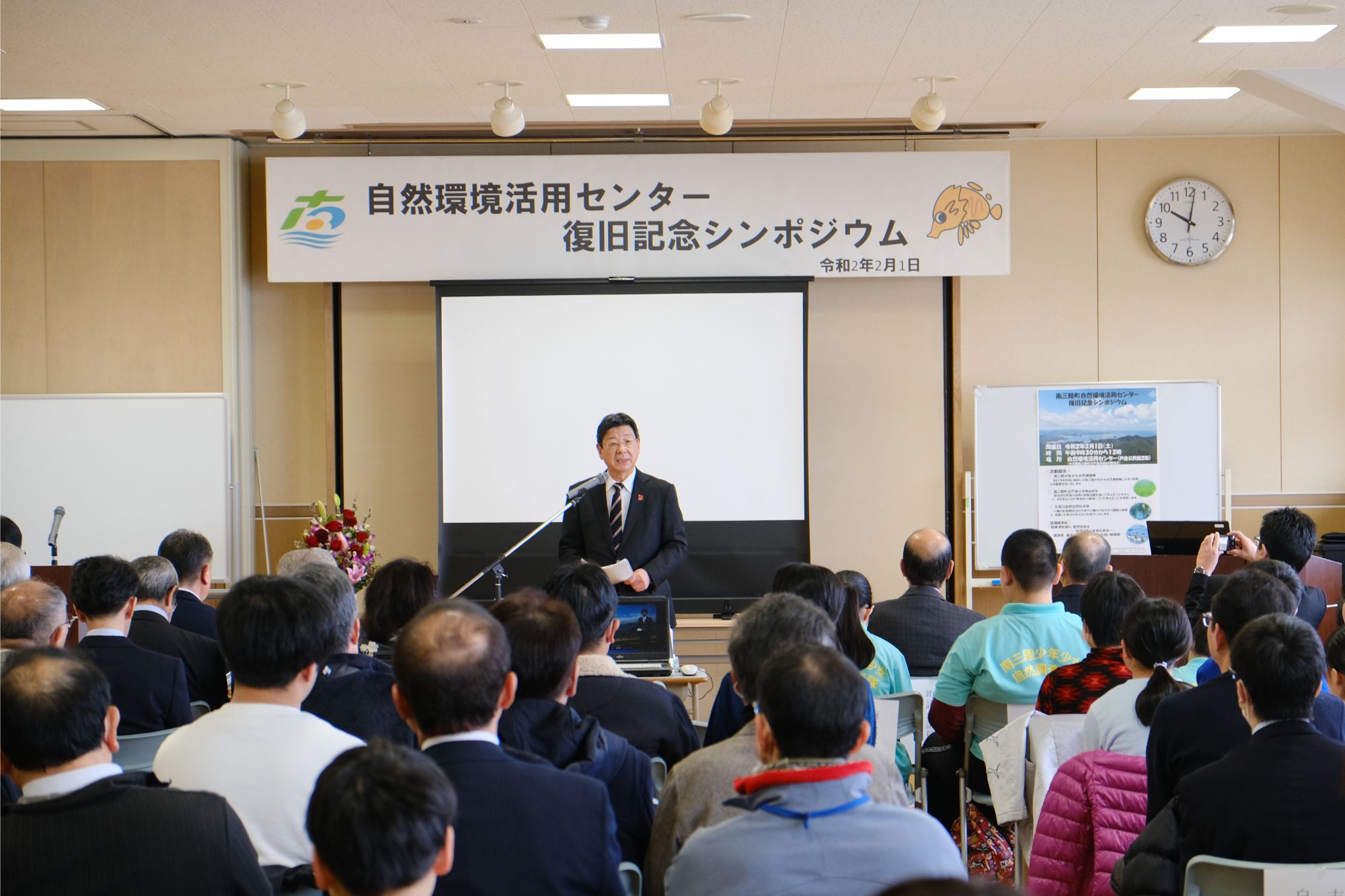 「自然環境活用センター 復旧記念シンポジウム」と書かれた横断幕が掲げられた壇上で阿部拓三さんが聴衆に向かって話している講演会の様子の写真