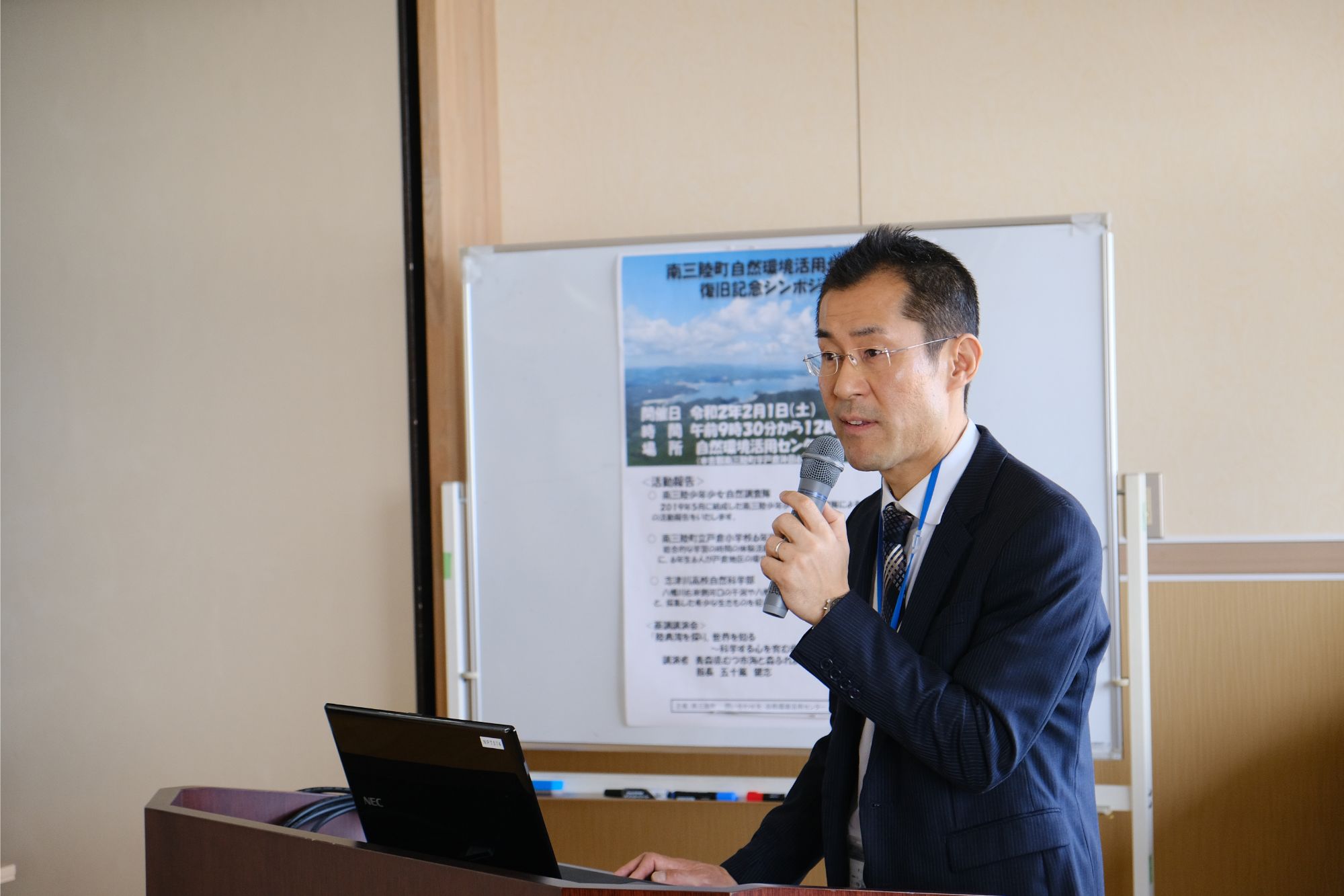 スーツ姿の人阿部拓三さんがマイクを持って壇上で話しており、背後にはポスターが掲示された白板がある講演の様子の写真