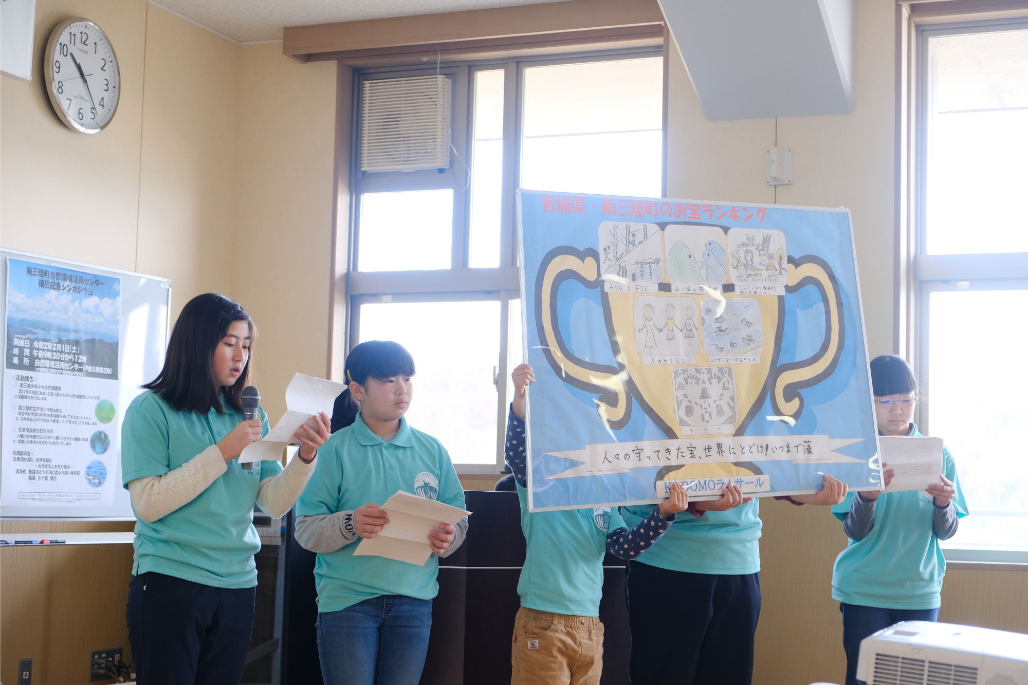 中央に立つ2人が優勝カップの様なイラストが描かれた大きなボードを持ち、左側の女の子が紙を持って発表を行っている様子の写真