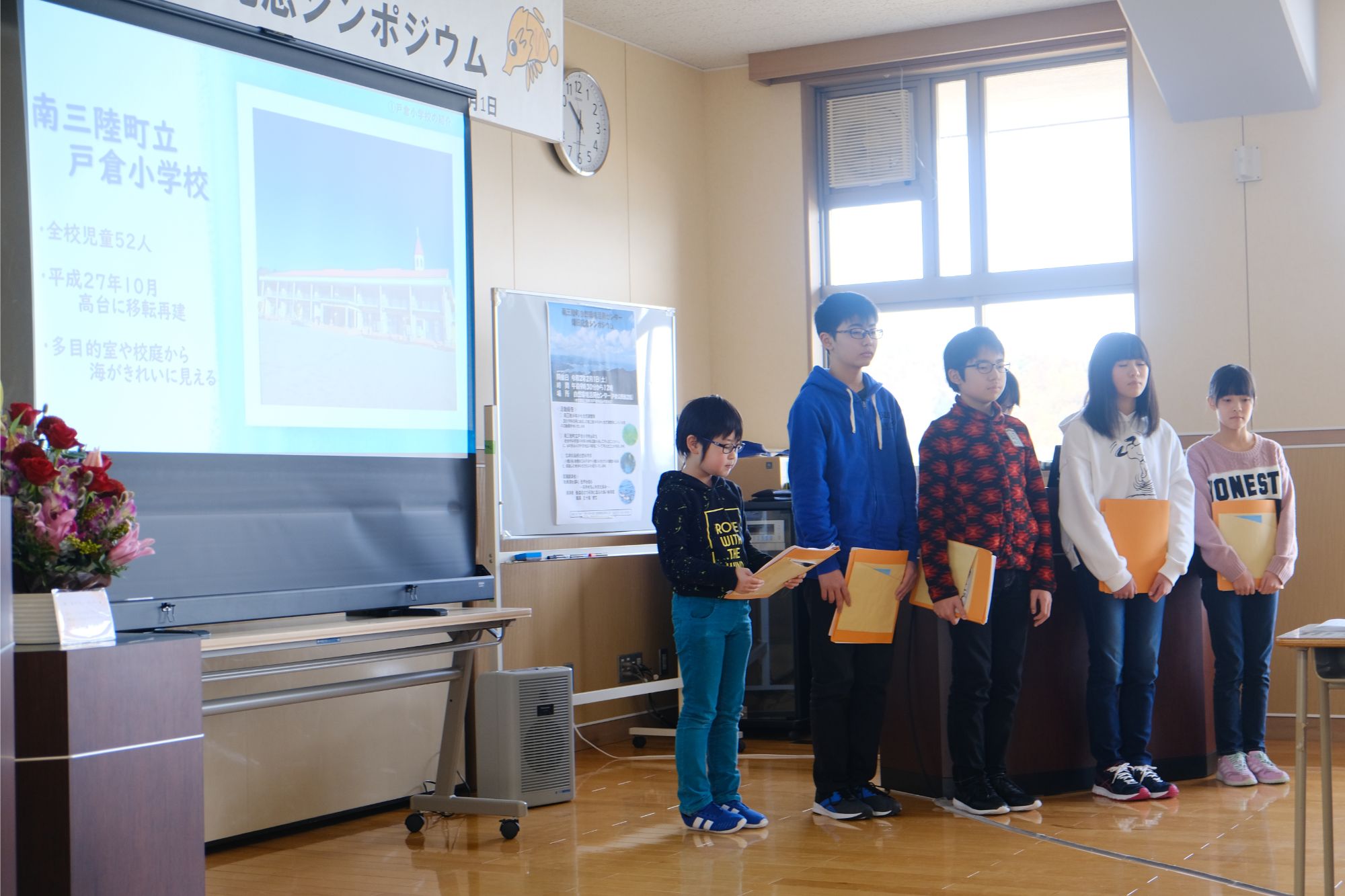 教室のような場所で5人の子どもたちが資料を手に発表しており、左側のスクリーンには「南三陸町立戸倉小学校」などの紹介文が映し出されている学校イベントの写真