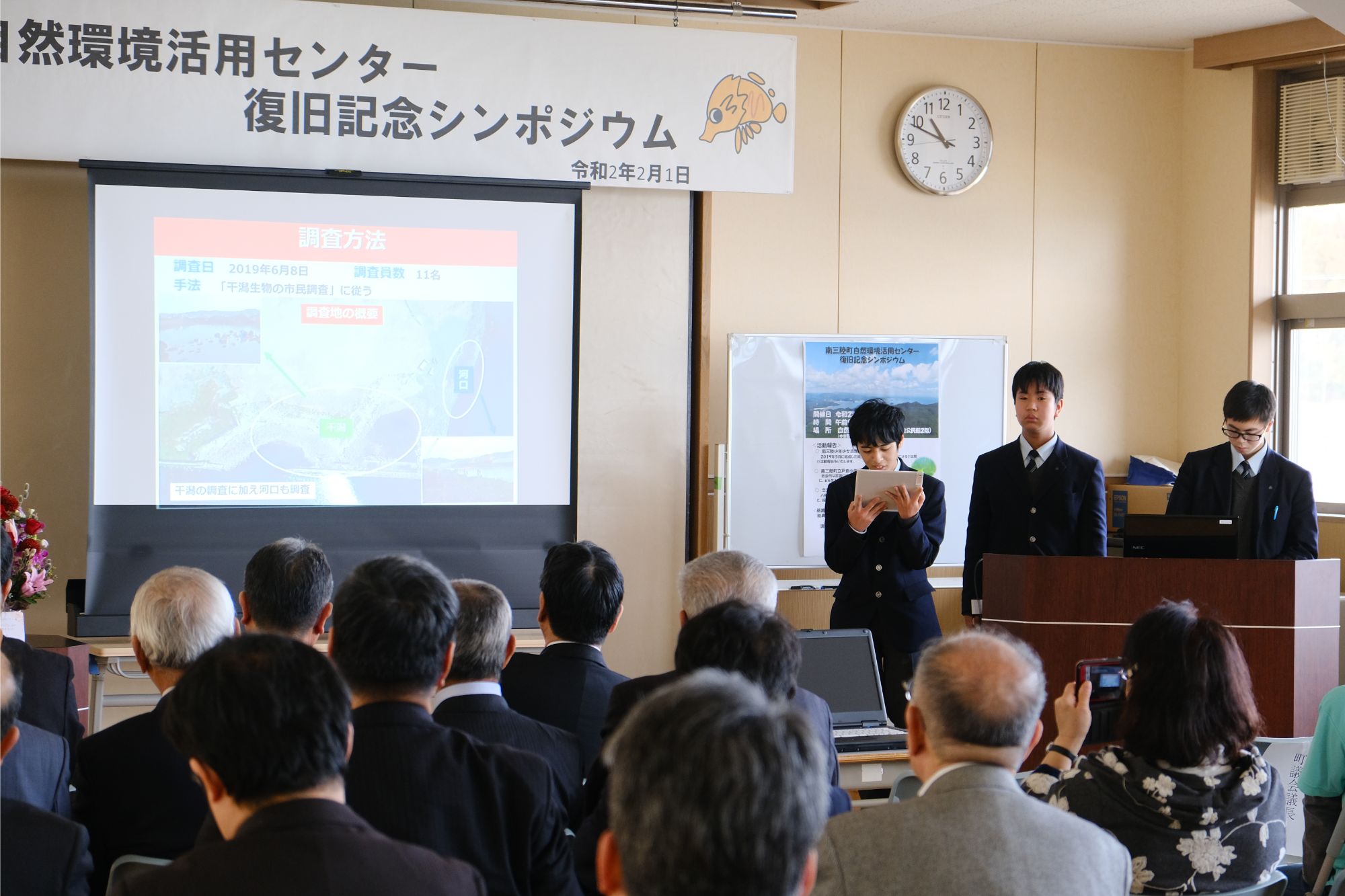 「自然環境活用センター 復旧記念シンポジウム」と書かれた横断幕の下で、3人の男子生徒の登壇者が発表を行い、聴衆がスクリーンに映されたスライドを見つめる中、発表を聞いている講演会の写真