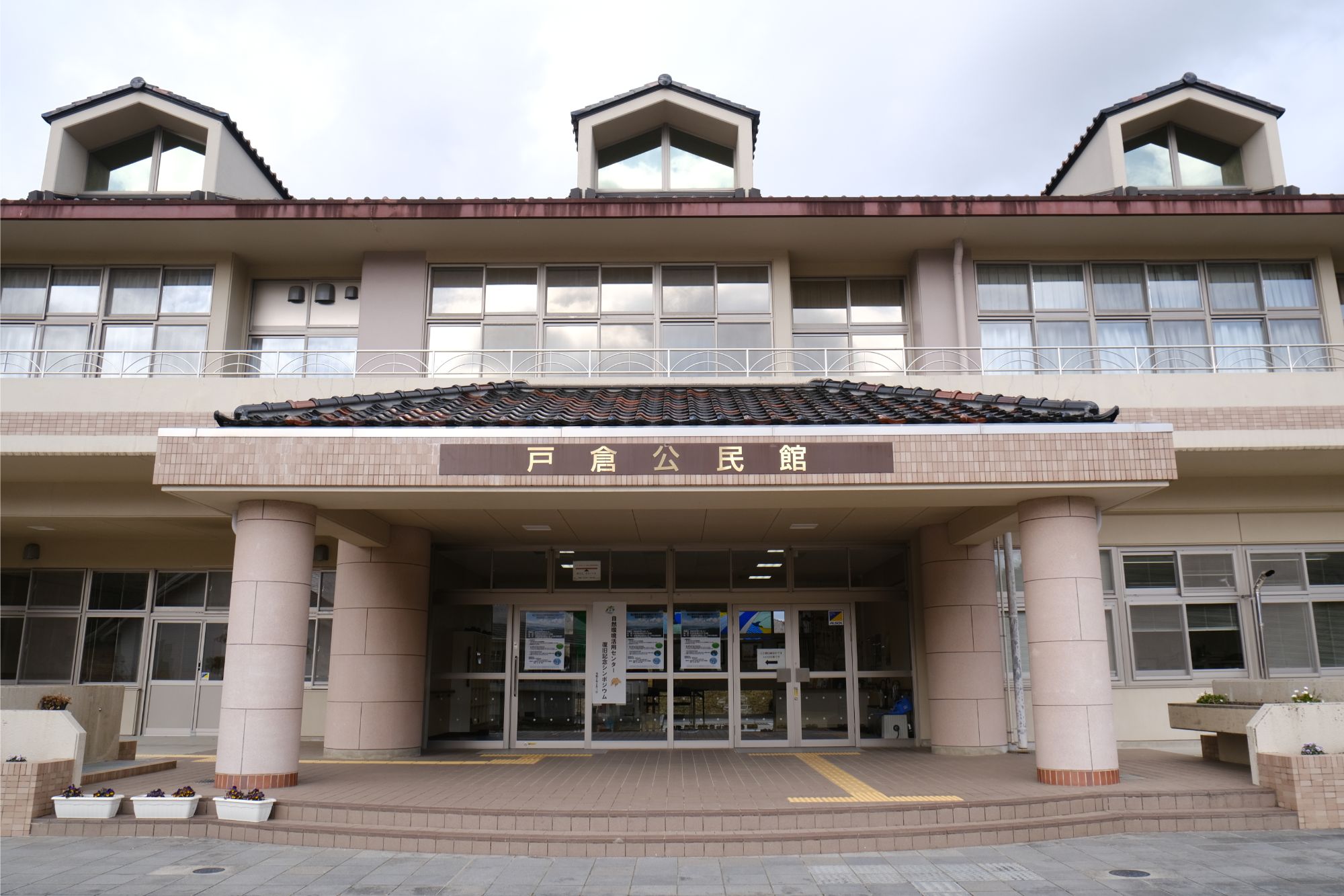 「戸倉公民館」と書かれた看板が掲げられた建物の外観で、左右対称のデザインに大きな窓と三つの特徴的な屋根があり、入口には二本の柱とガラス扉があり、扉や窓には掲示物が貼られている公共施設の写真