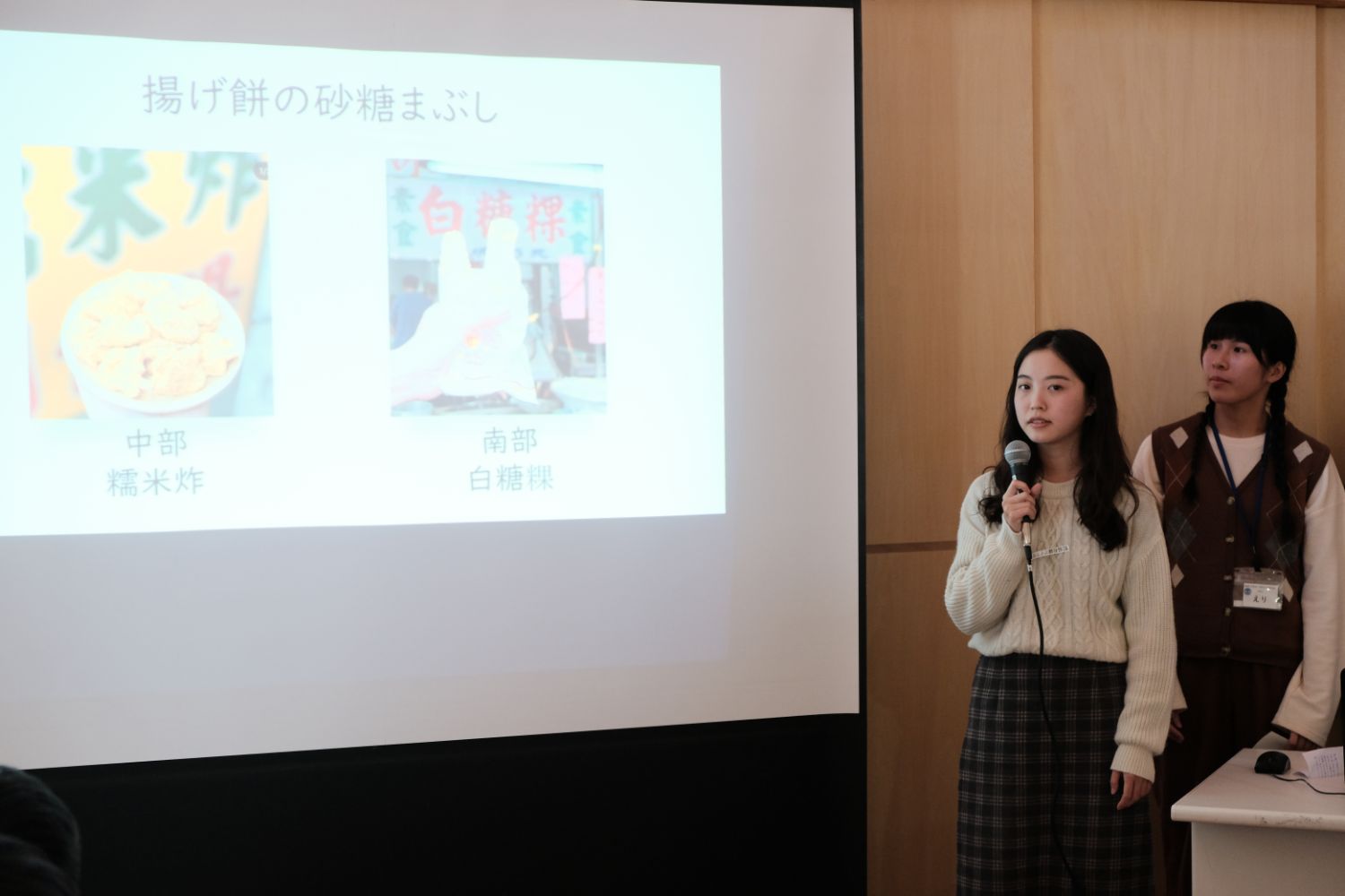 スクリーンに「揚げ餅の砂糖まぶし」と書かれ2枚の写真が表示されたスクリーンの右側に、マイクを持った台湾の女子大生が台湾のグルメについて発表を行っている写真