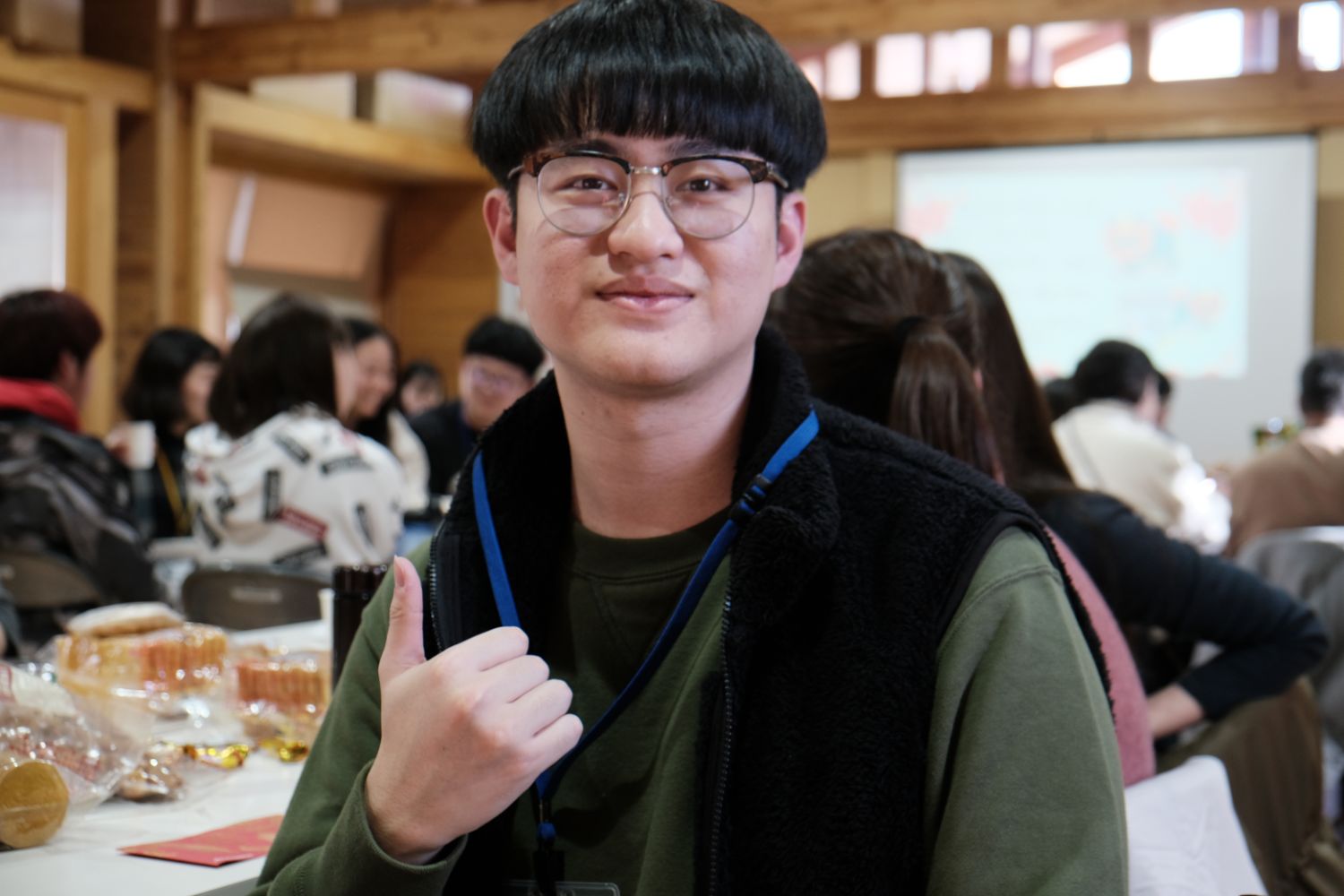 モスグリーン色の服を着た男子大生が、カメラに向かって右手でサムズアップのポーズをとっている写真