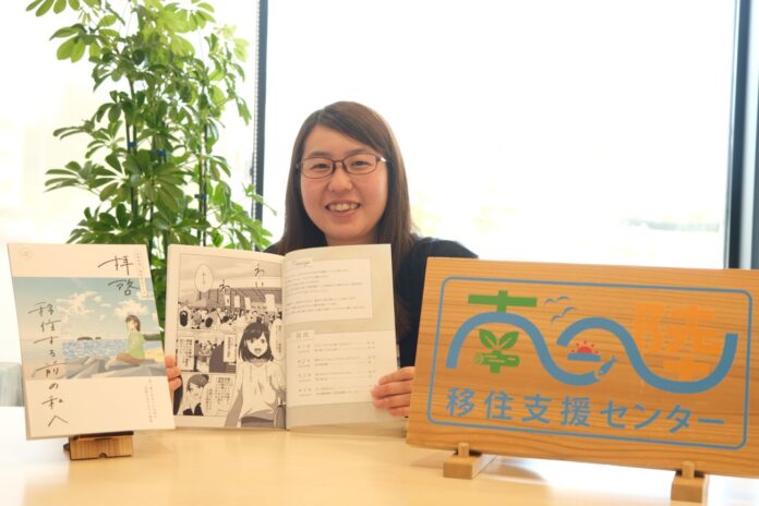 及川さんが笑顔で、移住ガイド冊子と漫画を広げて紹介しており、横には移住支援センターと書かれた木の看板が置かれている写真