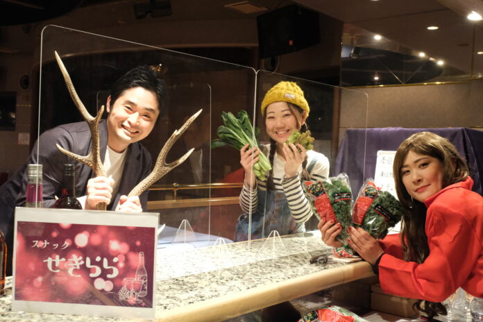 左の男性は笑顔でシカの角を両手に持ち、中央の女性はニット帽をかぶり、葉物野菜を両手に持って、右の女性は長い髪で赤い服を着ていて、複数の袋入りの葉物野菜を持っている写真