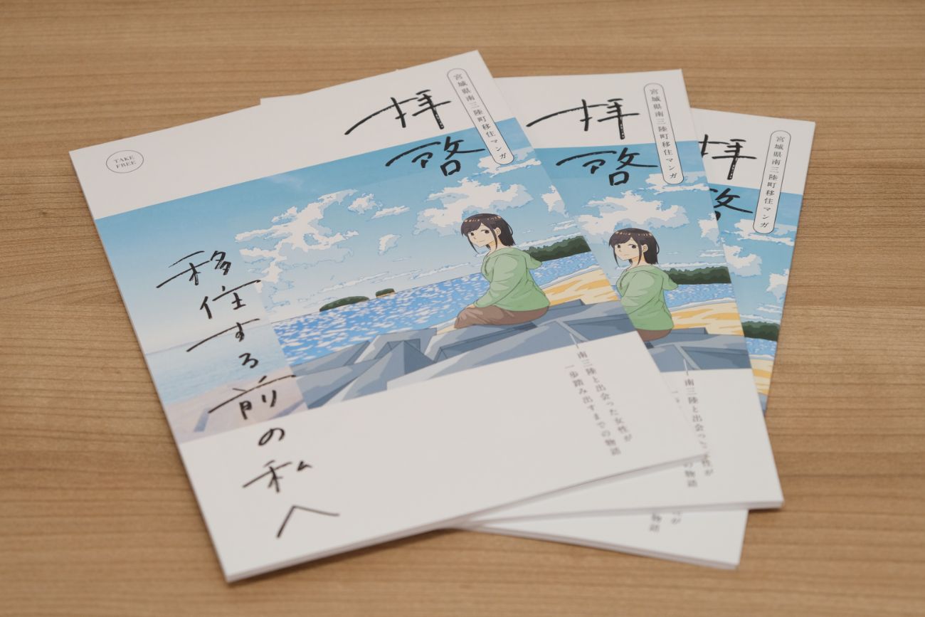 アニメ風に描かれた表紙の移住ガイド冊子がテーブルに3冊重ねて置かれており、南三陸町の海辺に座る女性のイラストが描かれている写真