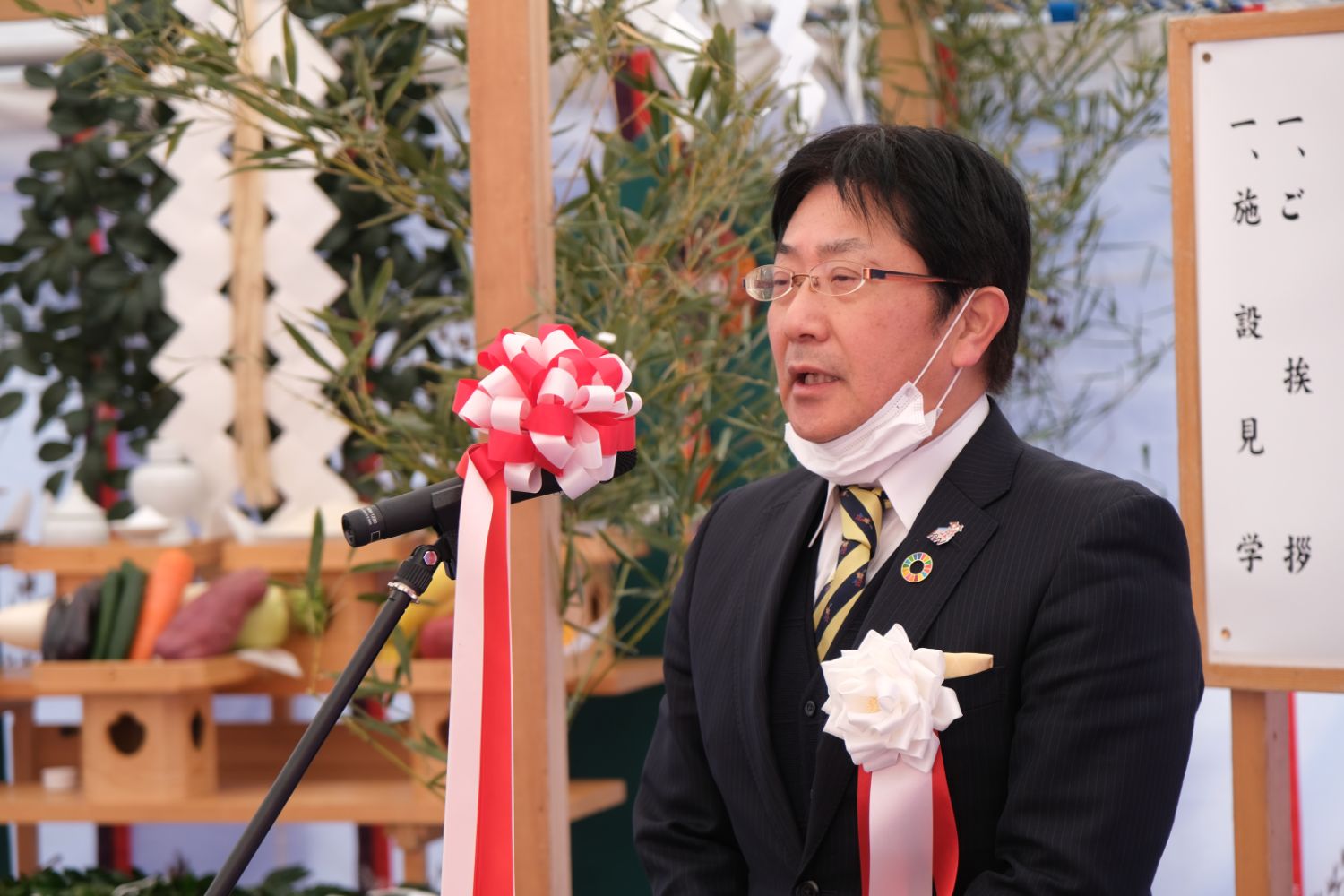 背後には榊や野菜が供えられた神棚が並び、紅白のリボンが付いたマイクの前で、白い胸章をつけた松本俊人さんが地鎮祭の祭壇の前で挨拶を述べている写真。