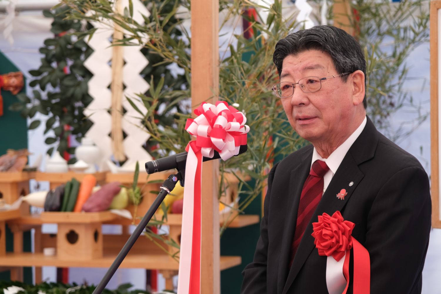 背後には榊や野菜が供えられた神棚が並び、紅白のリボンが付いたマイクの前で、赤い胸章をつけた佐藤仁町長が地鎮祭の祭壇の前で挨拶を述べている写真。