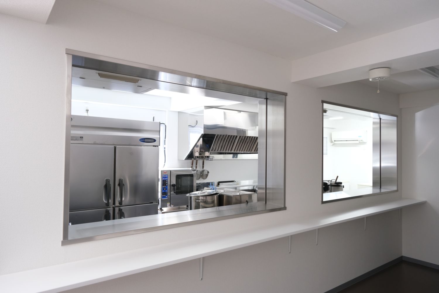 白い壁と天井に囲まれた清潔感のある室内に、大型冷蔵庫やコンロ、換気フードなどの厨房設備が整い、手前のカウンター越しにキッチン全体が見渡せる給食室の写真。