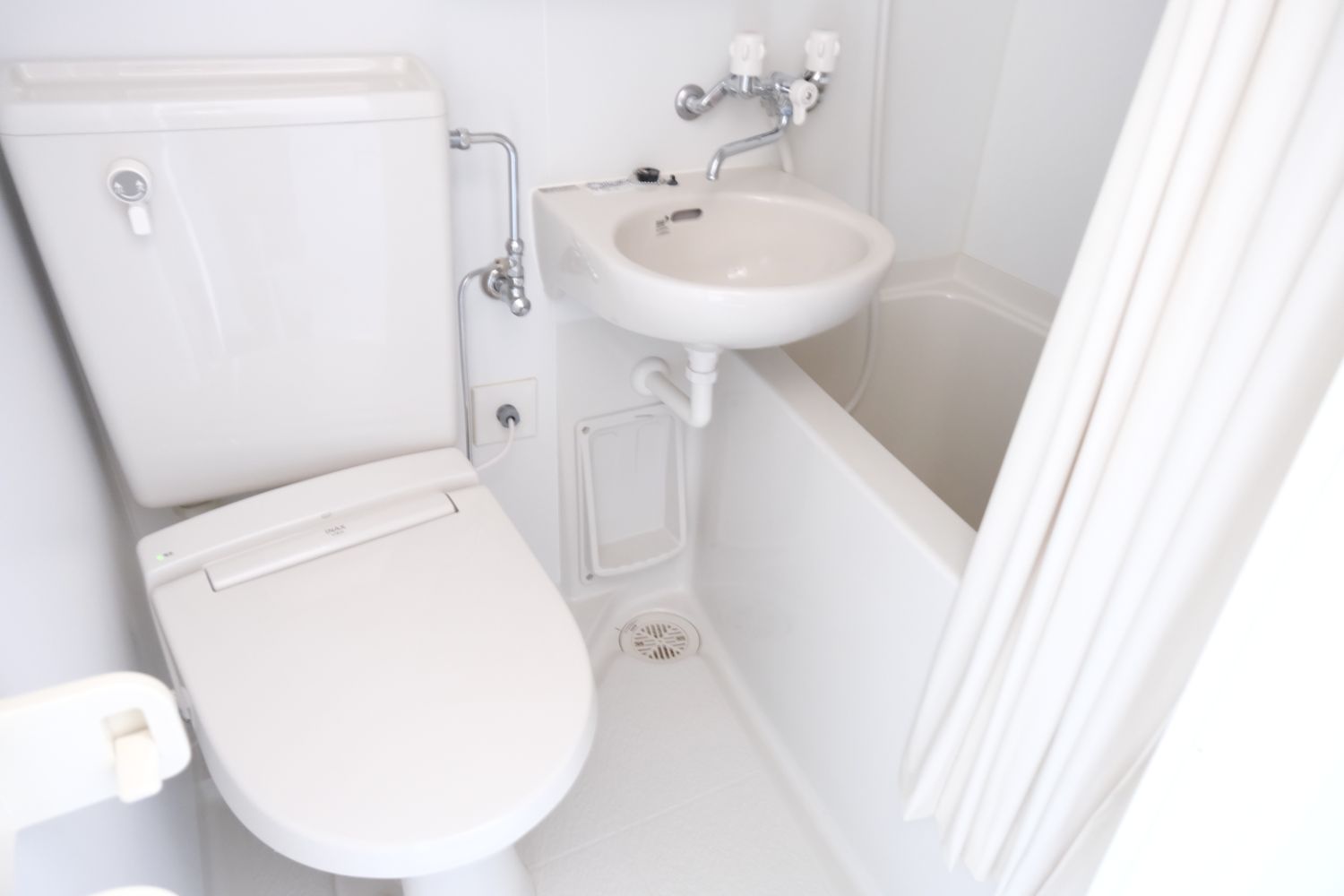 白い壁に囲まれたユニットバスの空間に、洋式トイレと小さな洗面台、カーテンで仕切られた浴槽が配置された清潔感のあるバスルームの写真。