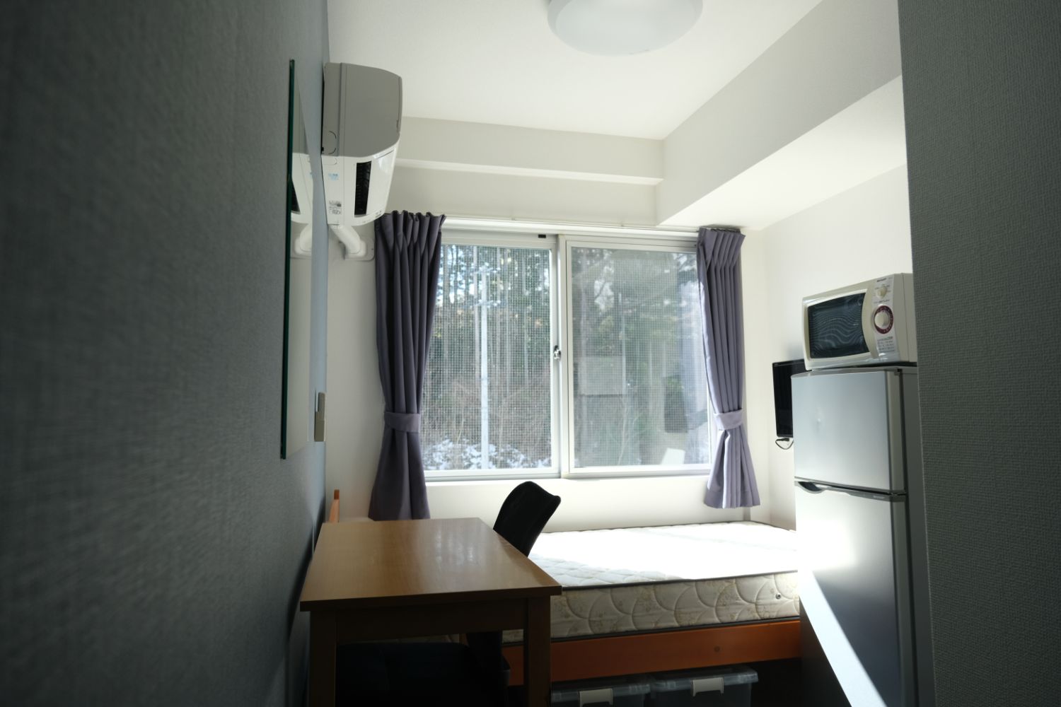 自然光が差し込む明るい室内にシングルベッドと木の机、冷蔵庫と電子レンジが備え付けられた細長いワンルームの部屋の写真。