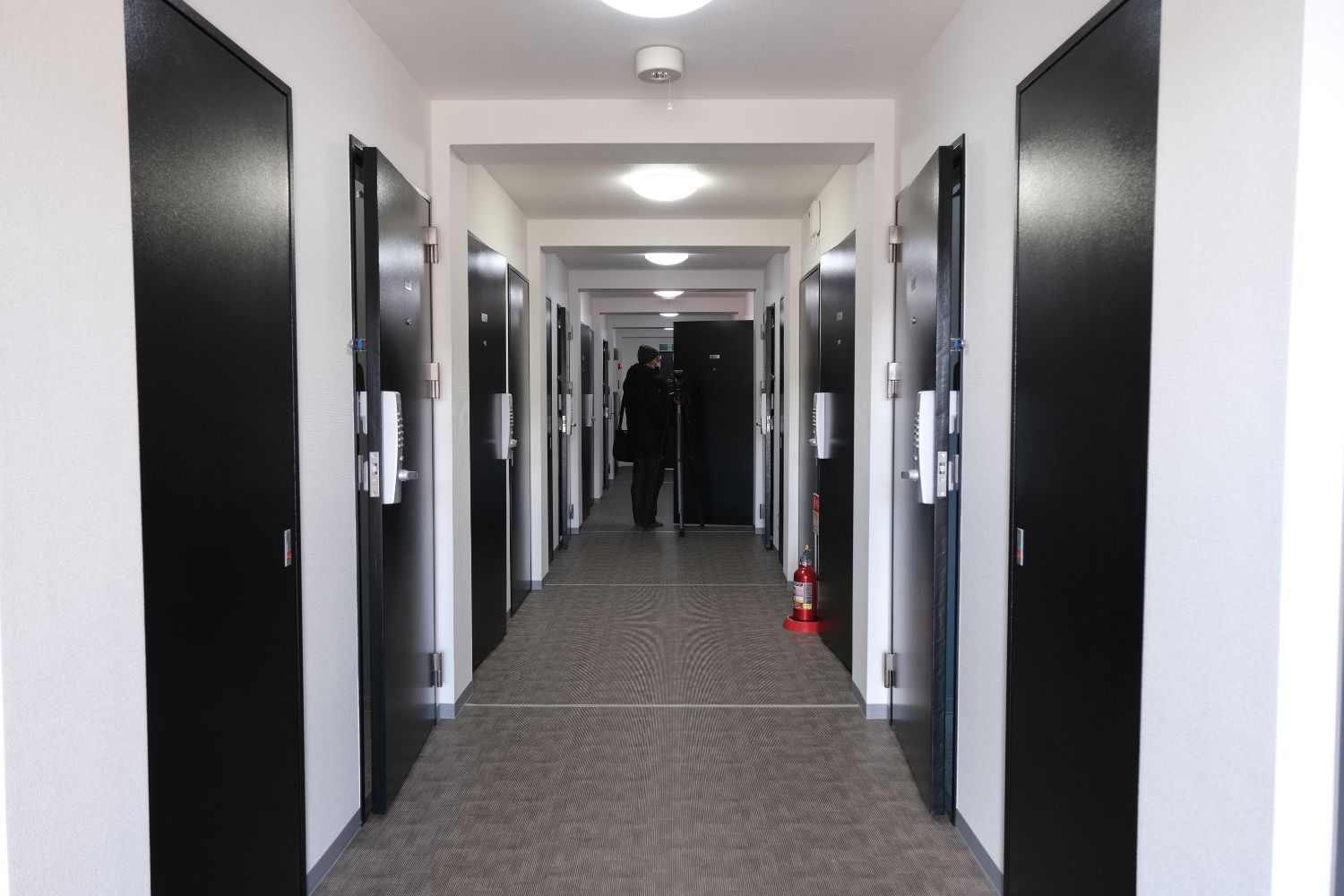 白い壁と黒い扉が左右に並ぶ明るい屋内廊下の奥に人物が立ち、天井照明が等間隔に設置された学生寮の廊下の写真。