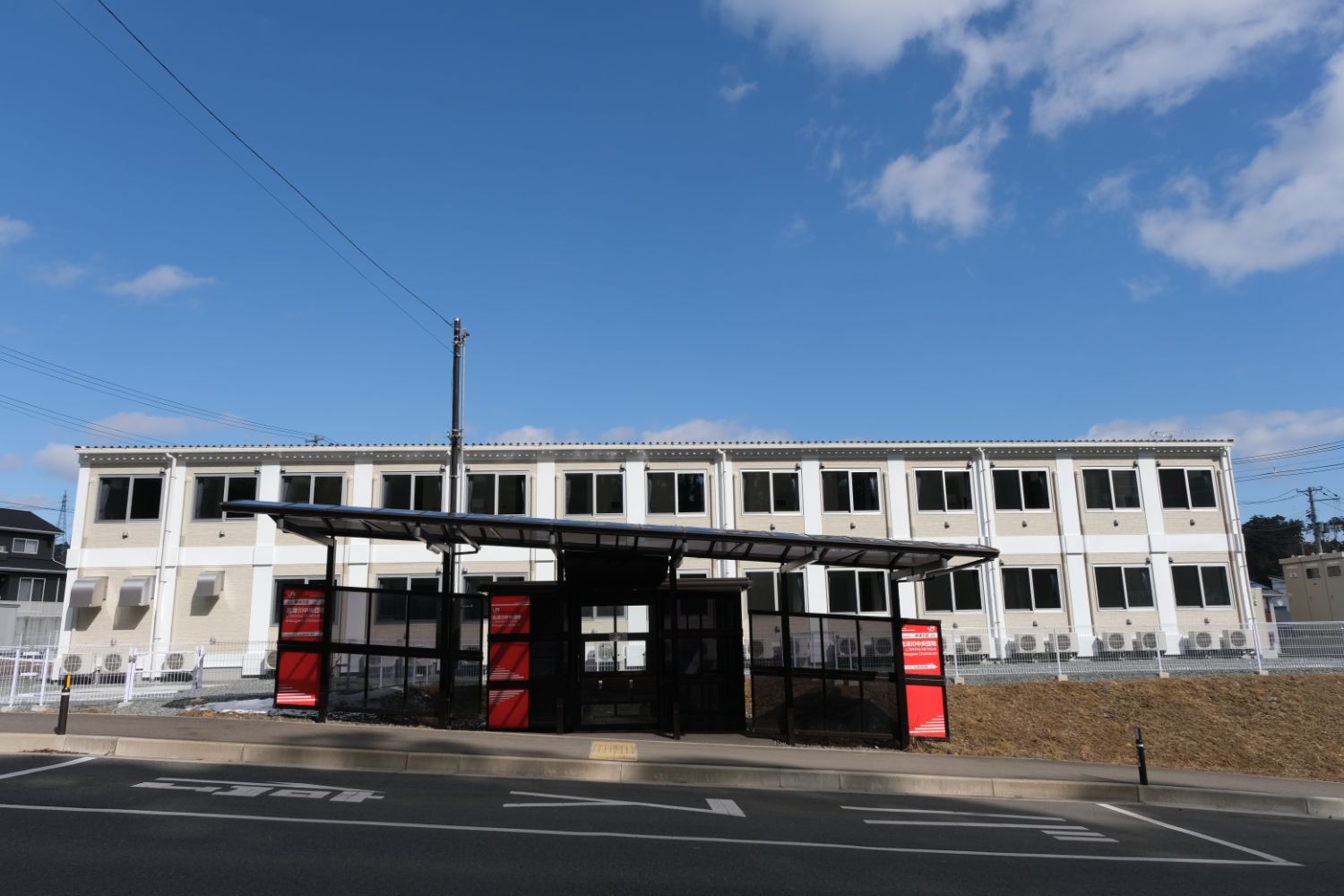 白とベージュの二階建ての建物が奥に建ち、手前には黒い屋根と赤い案内板のあるガラス張りのバス停のような構造物が道路沿いに設置されている、施設外観の写真。