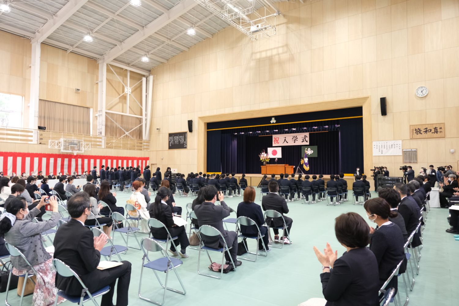 体育館内で新入生や保護者が整列して席に着き、壇上に日の丸と「祝 入学式」の垂れ幕が掲げられた入学式の様子の写真