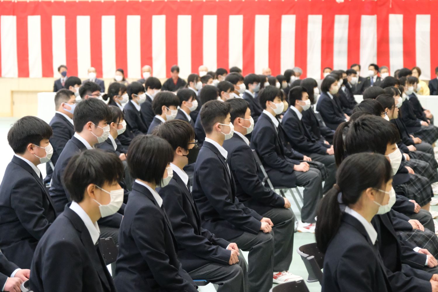 マスクを着用した新入生たちが整然と座り、紅白幕が貼られた体育館での入学式に参加している様子の写真