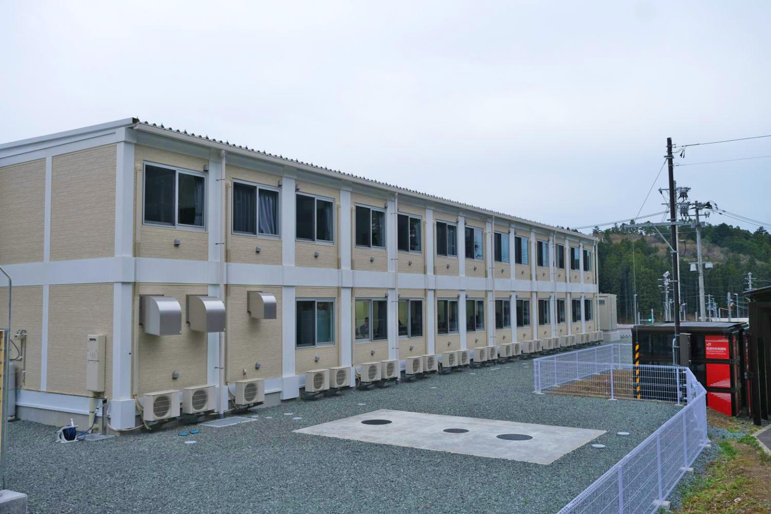 白いフェンスで囲まれた砂利の敷地内に建つベージュに白のラインが入った2階建ての四角い建物の外にエアコンの室外機がたくさん並んでいる寮の外観写真