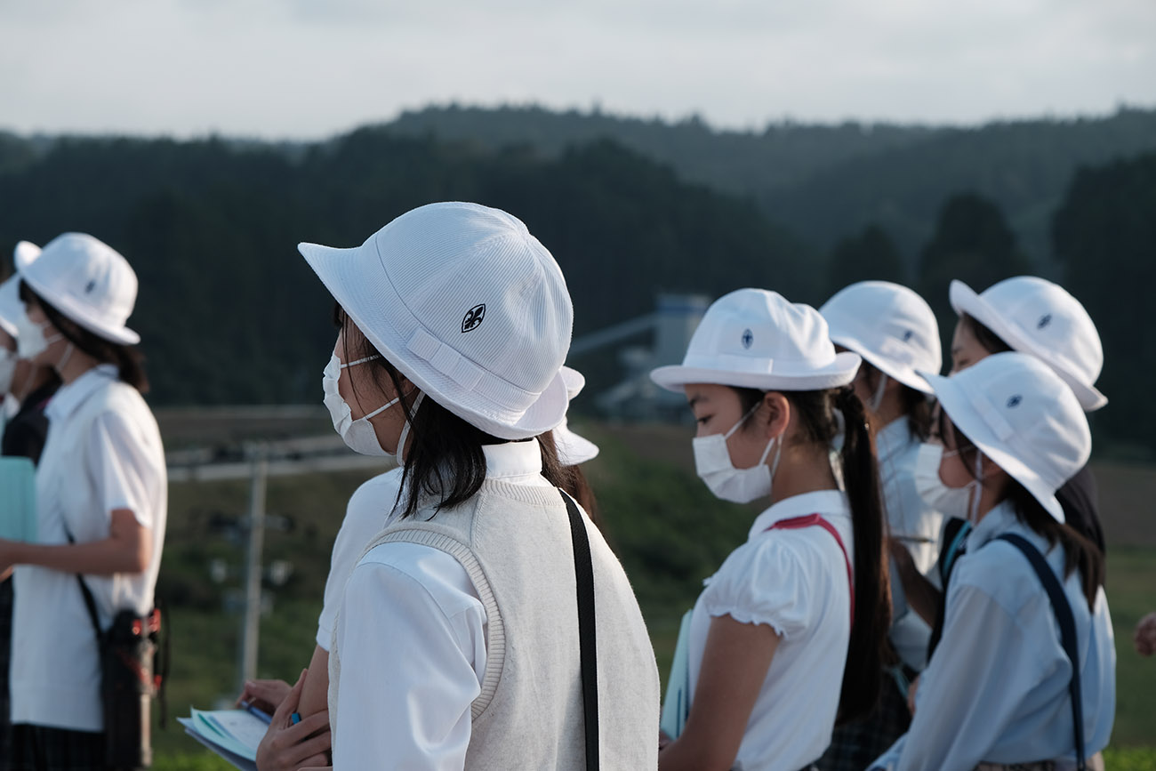 白い帽子とマスクを着用した女子学生たちが、ノートを手にして真剣な表情で話を聞いている写真