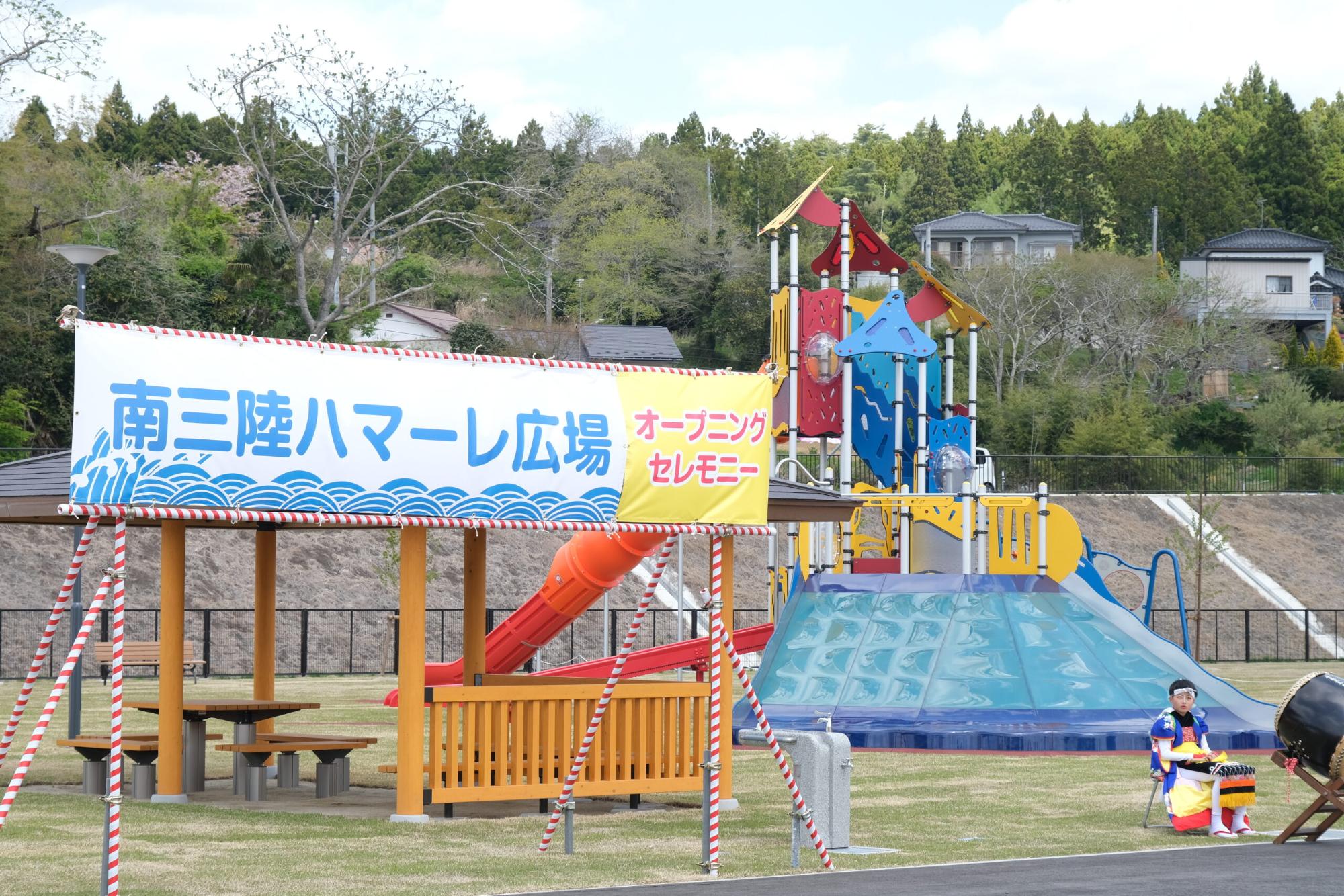 「南三陸ハマーレ広場 オープニングセレモニー」と書かれた横断幕の後ろに東屋とカラフルな大型遊具がある写真