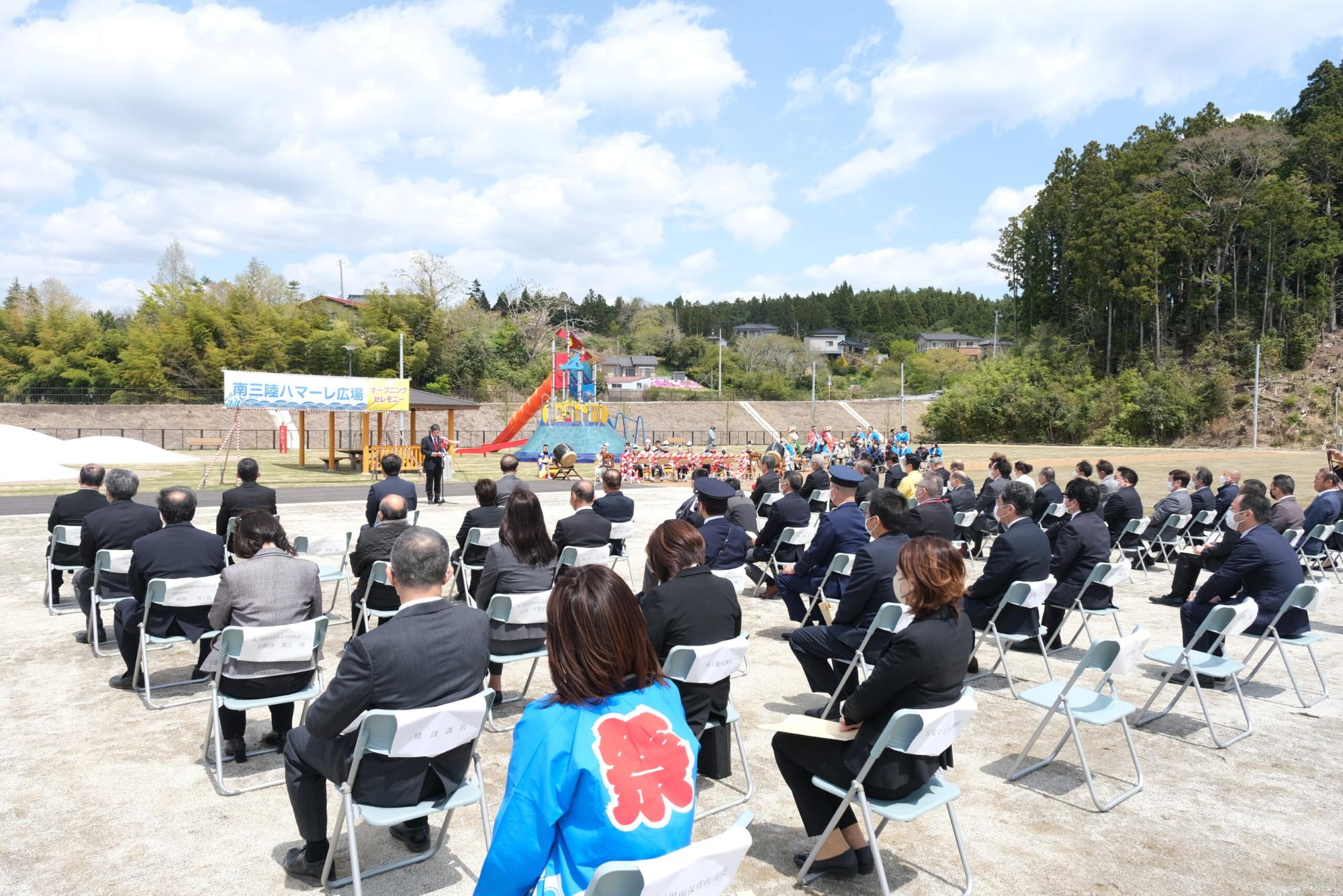 カラフルな大型遊具がある広場を背に男性が立ち、広場の方を向いて並んだ椅子に大勢の関係者たちが座っているセレモニーの様子の写真