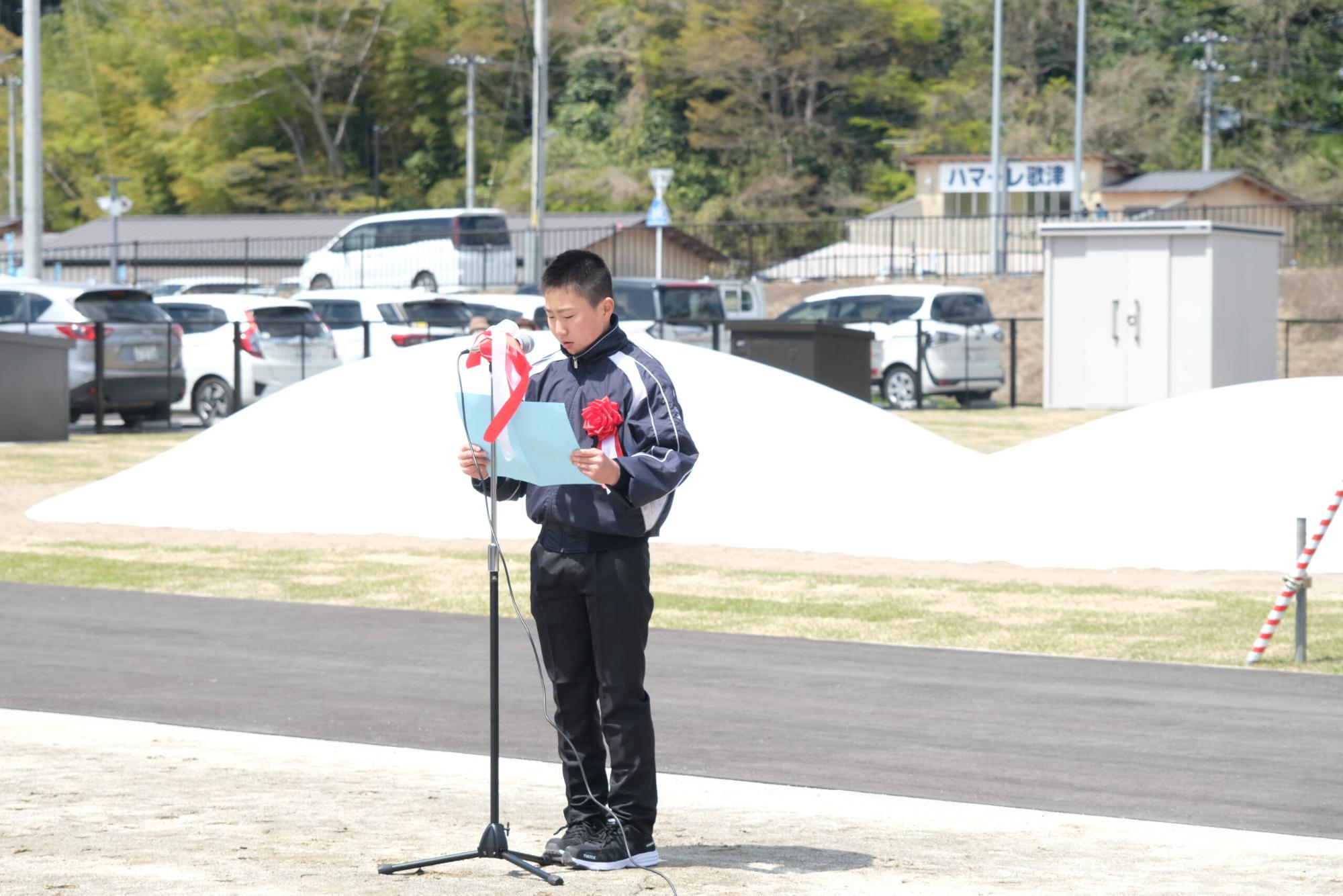 胸に赤いリボンをつけた千葉さんが、マイクの前に立ち、青い紙を読みながら話している写真